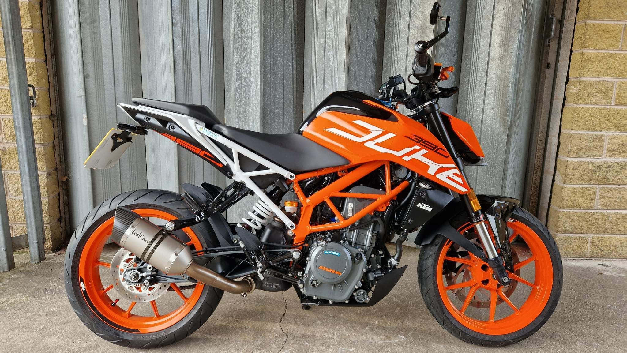 橙色KTM Duke 390機車圖片 壁紙 — 一輛鮮豔的橙色KTM Duke電單車停泊在波紋金屬和磚牆旁。