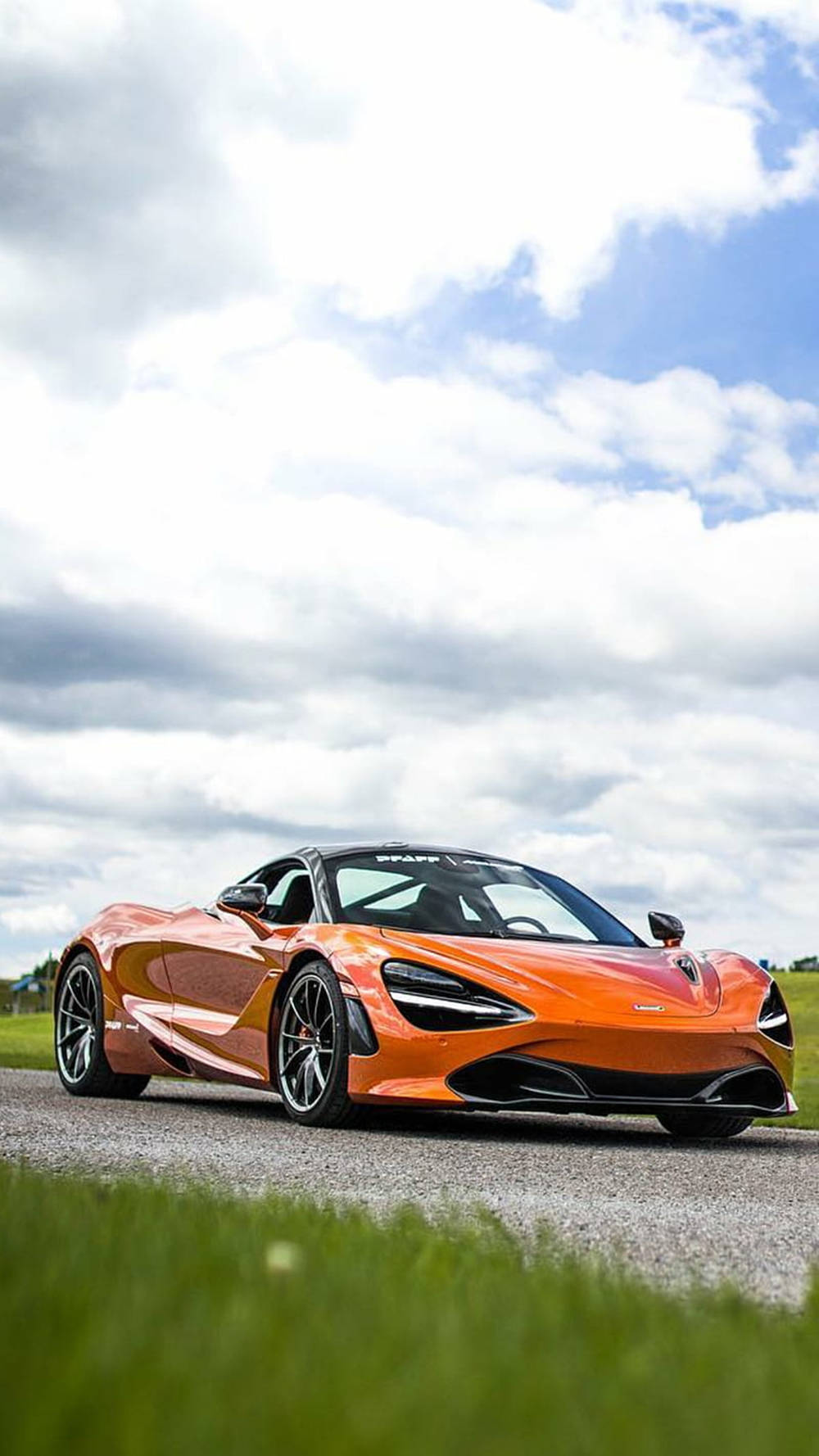 橙色McLaren 720S跑車在碎石路上，有草和多雲天空。