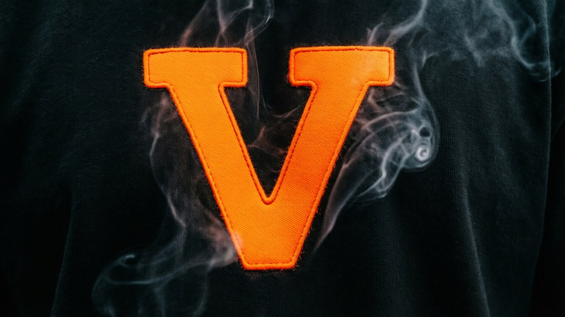 橙色V對黑 壁紙 — 黑色布料上一個大膽的橙色刺繡字母 V，周圍有白色煙霧旋轉。