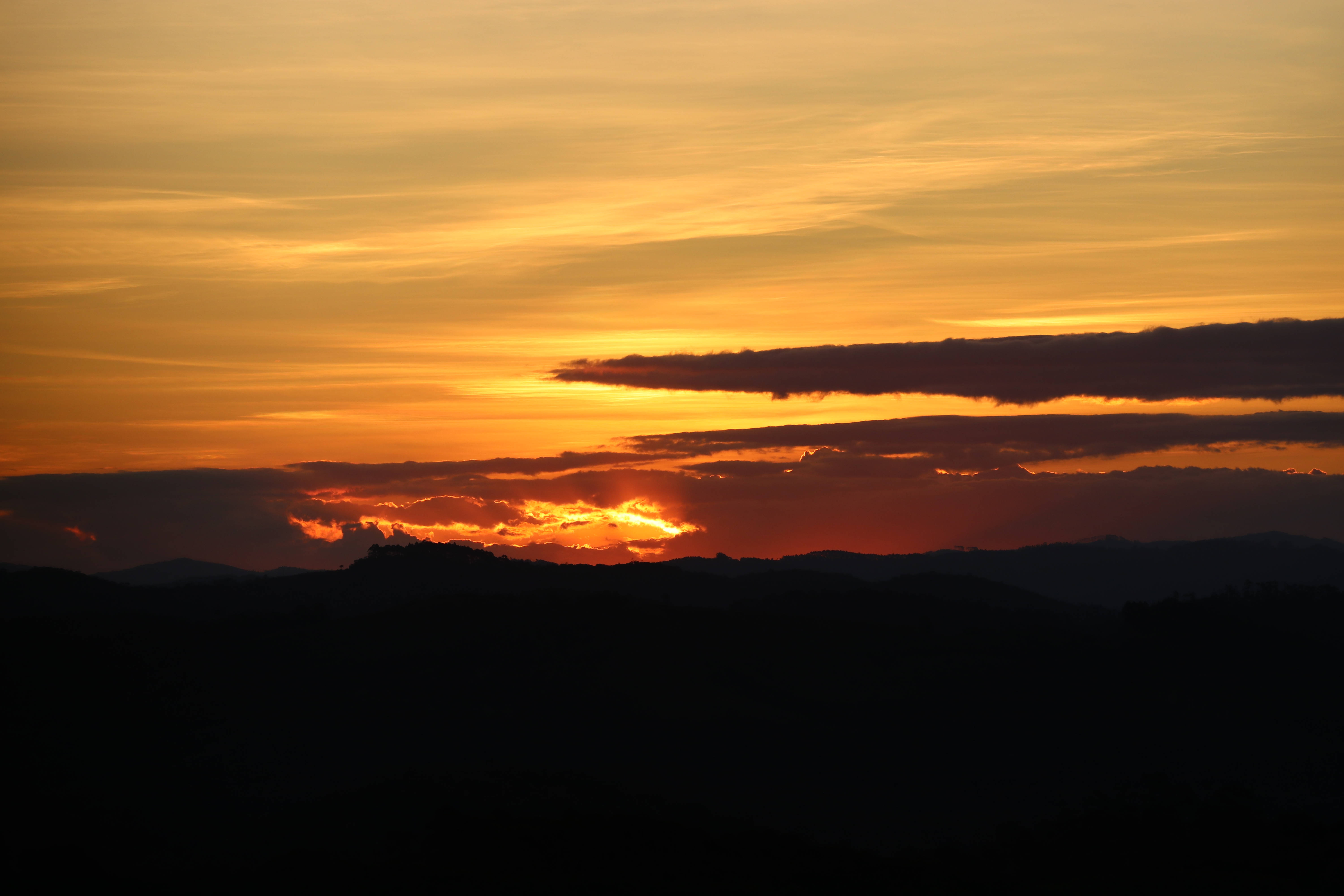 橙色天空與雲彩最佳桌面 壁紙 — 熾熱的夕陽將天空染成鮮豔的橙色和金色，雲朵在遠處山脈的襯托下形成剪影。