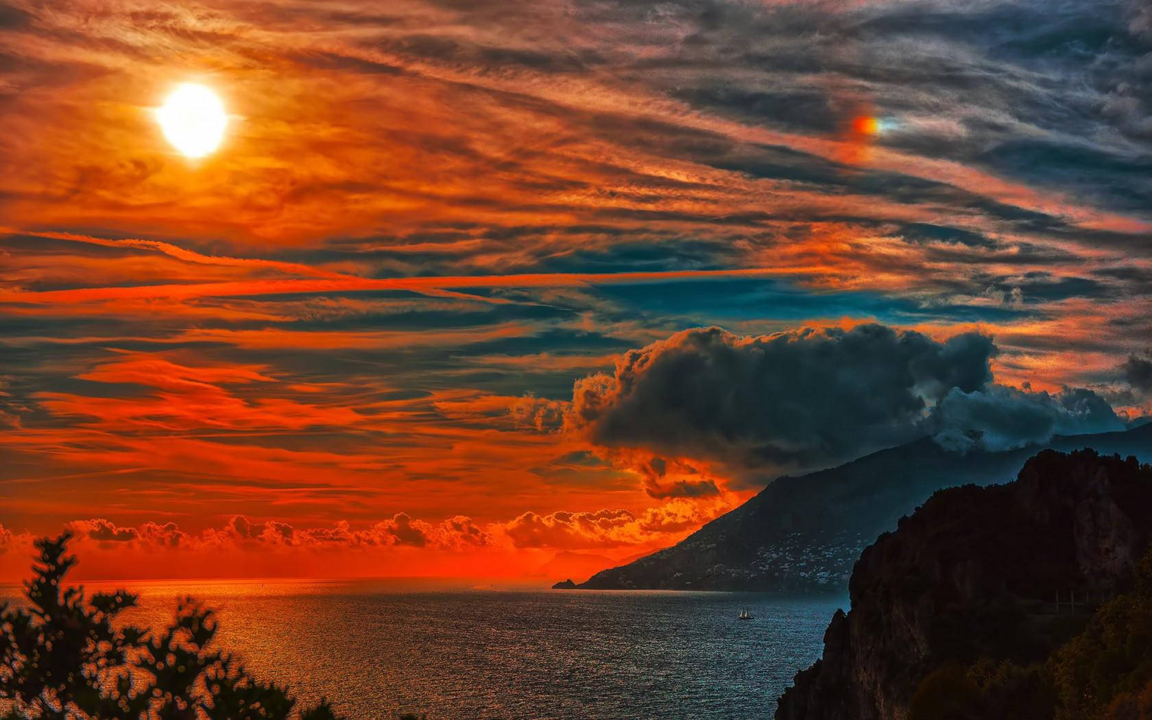 橙色日落天空高清 壁紙 — 熾熱的夕陽在平靜的海面上，以剪影的山脈和雲朵為背景，將天空塗成鮮豔的橙色和紅色色調。