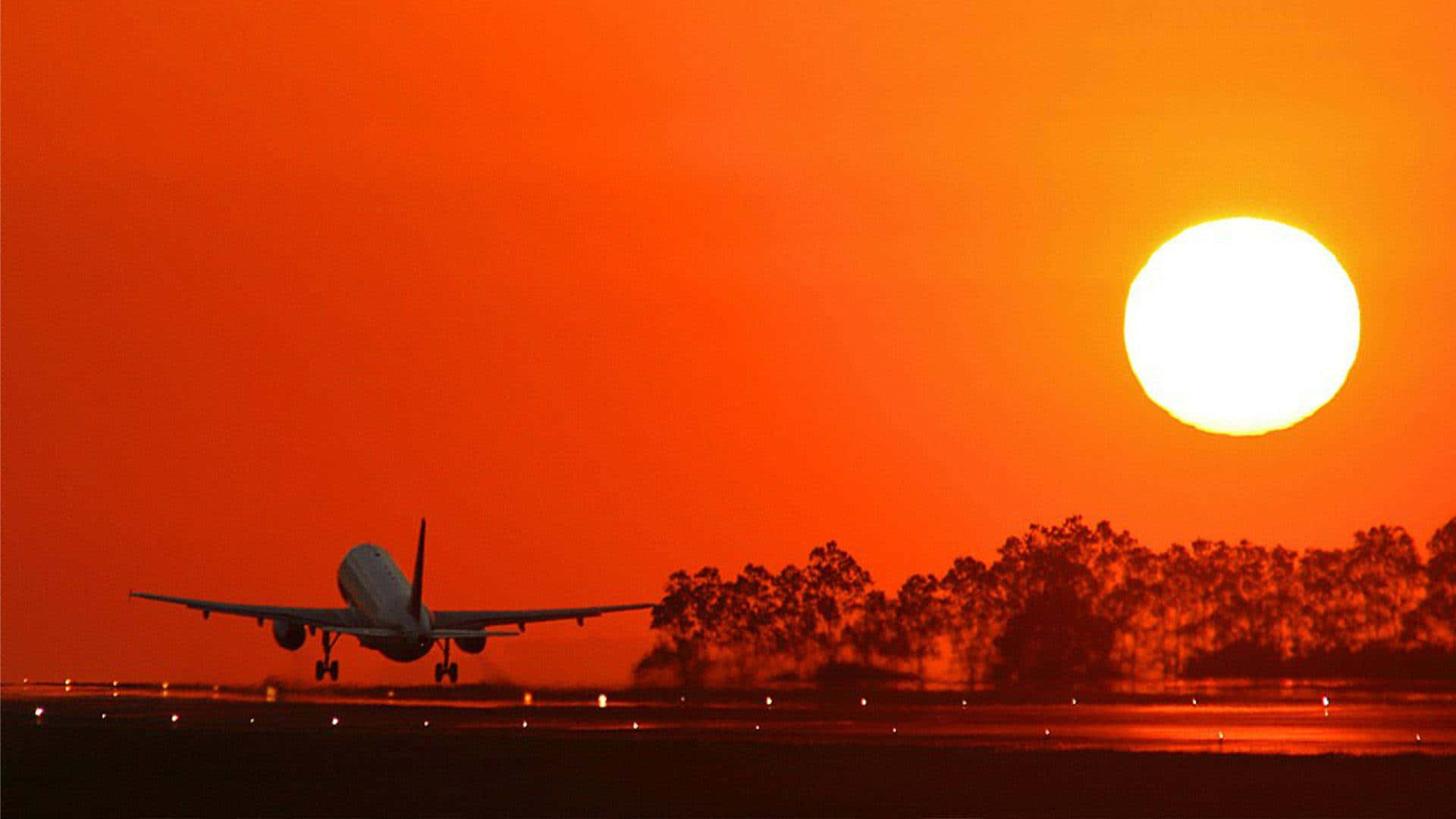 橙色日落與飛機背景 壁紙 — 一架飛機在火紅的橙色夕陽、發光的太陽和剪影樹木中下降。