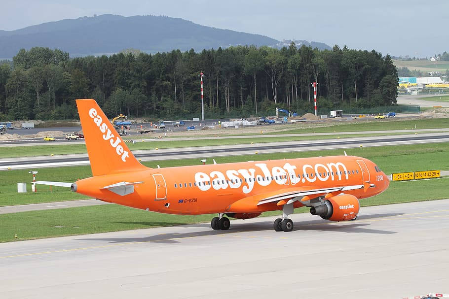橙色 easyJet 飛機喺機場停機坪上，後面有樹林同山丘。