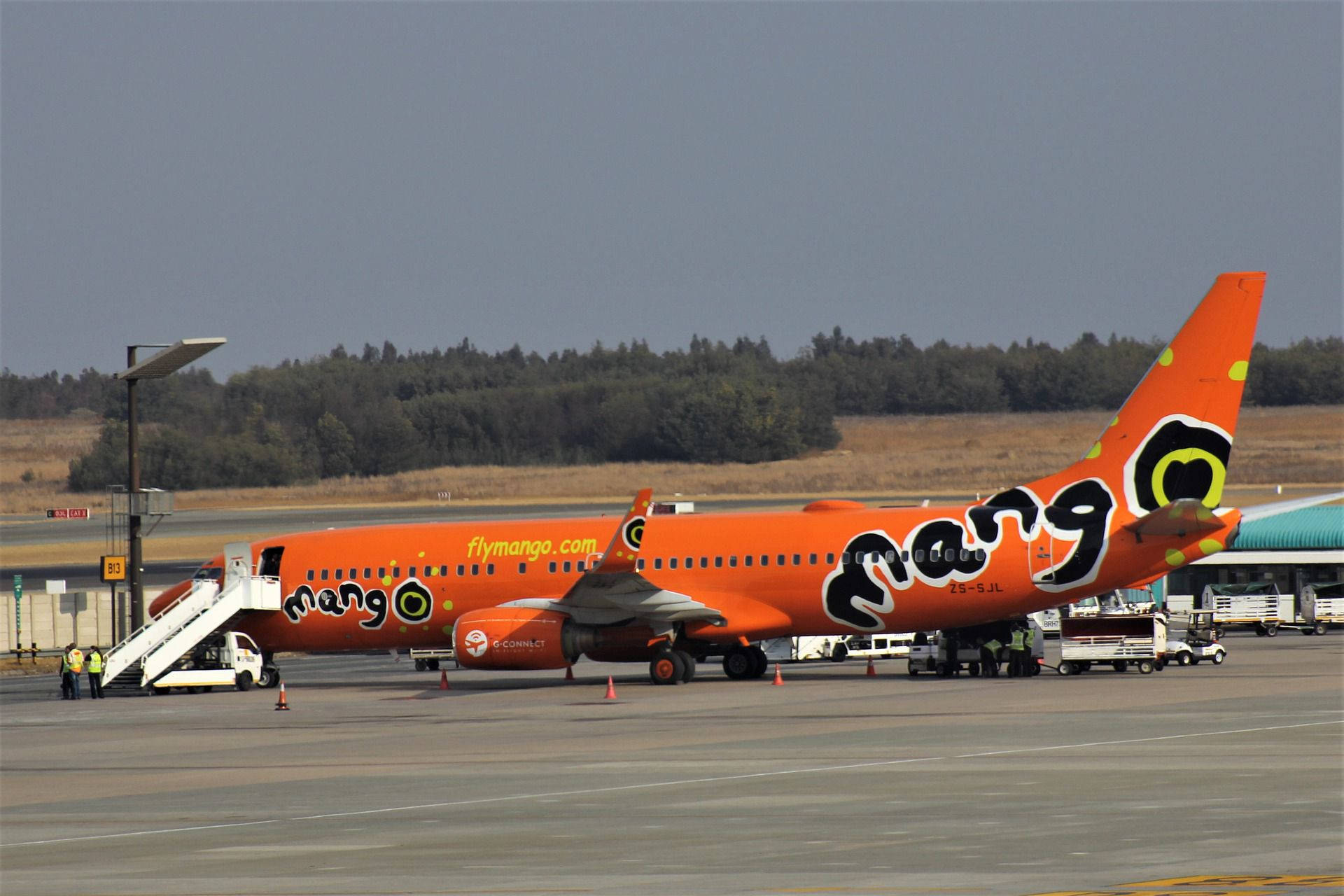 橙色Mango Airlines噴射機停在機場閘口，附有登機梯和地勤人員。