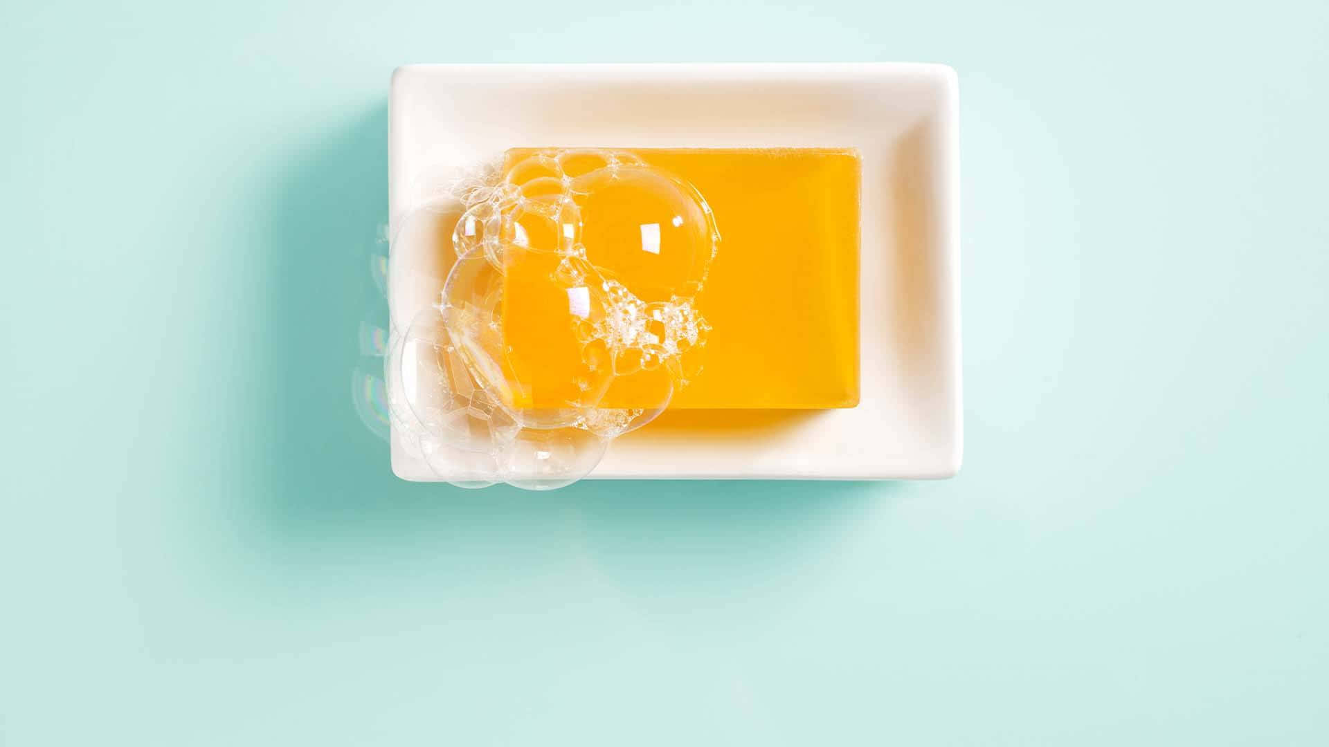 橙色香皂 壁紙 — 一塊鮮黃色肥皂帶有清澈氣泡，放在白色碟中，背景是柔和的薄荷綠色。