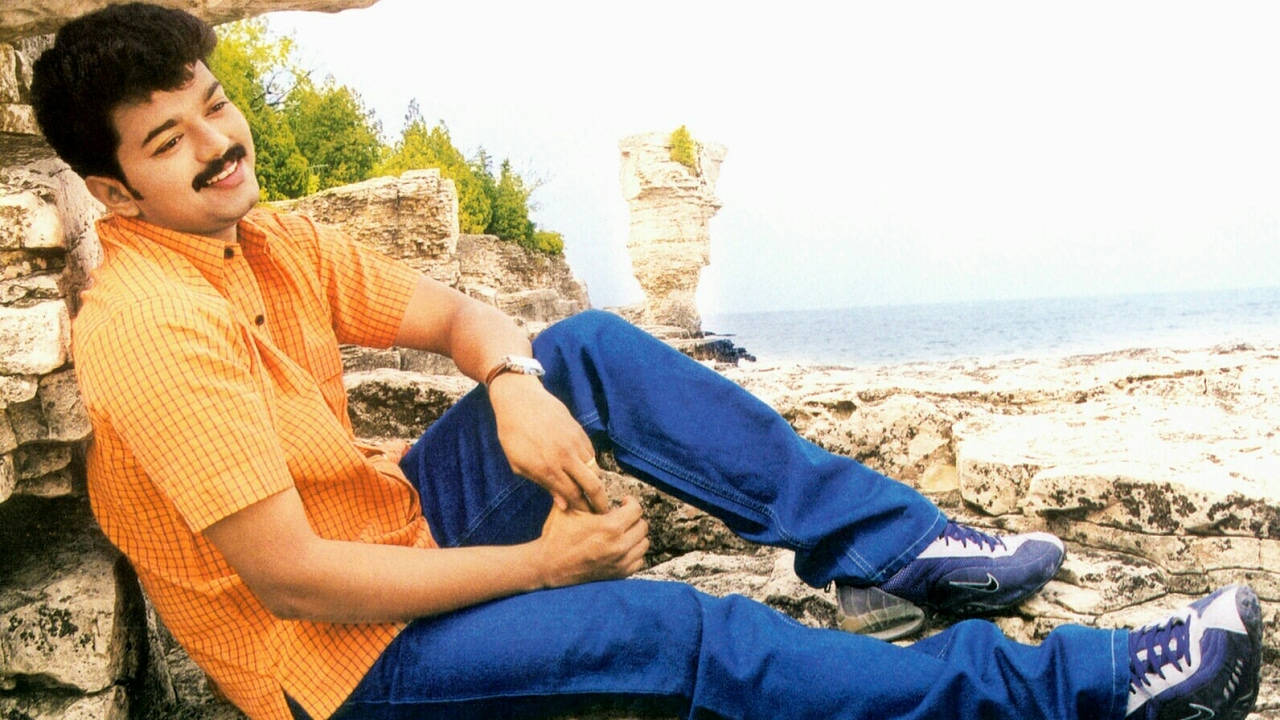穿著橙色襯衫和藍色牛仔褲的男人在岩石海邊峭壁上微笑。