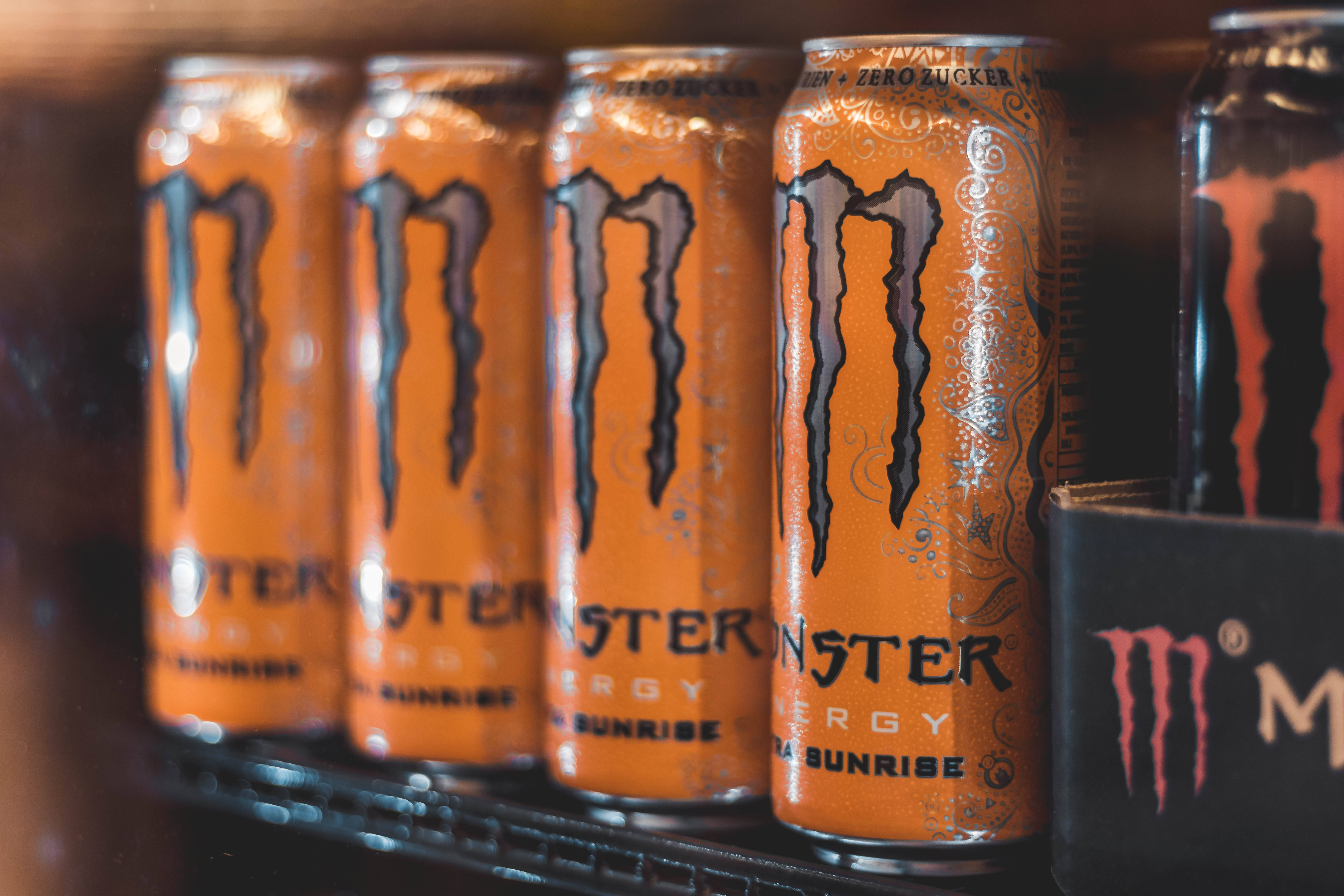 橙黑配色 Monster Energy 電解質飲料罐 壁紙 — 鮮橙色Monster Energy罐在深色架子上排列的特寫，背景模糊。