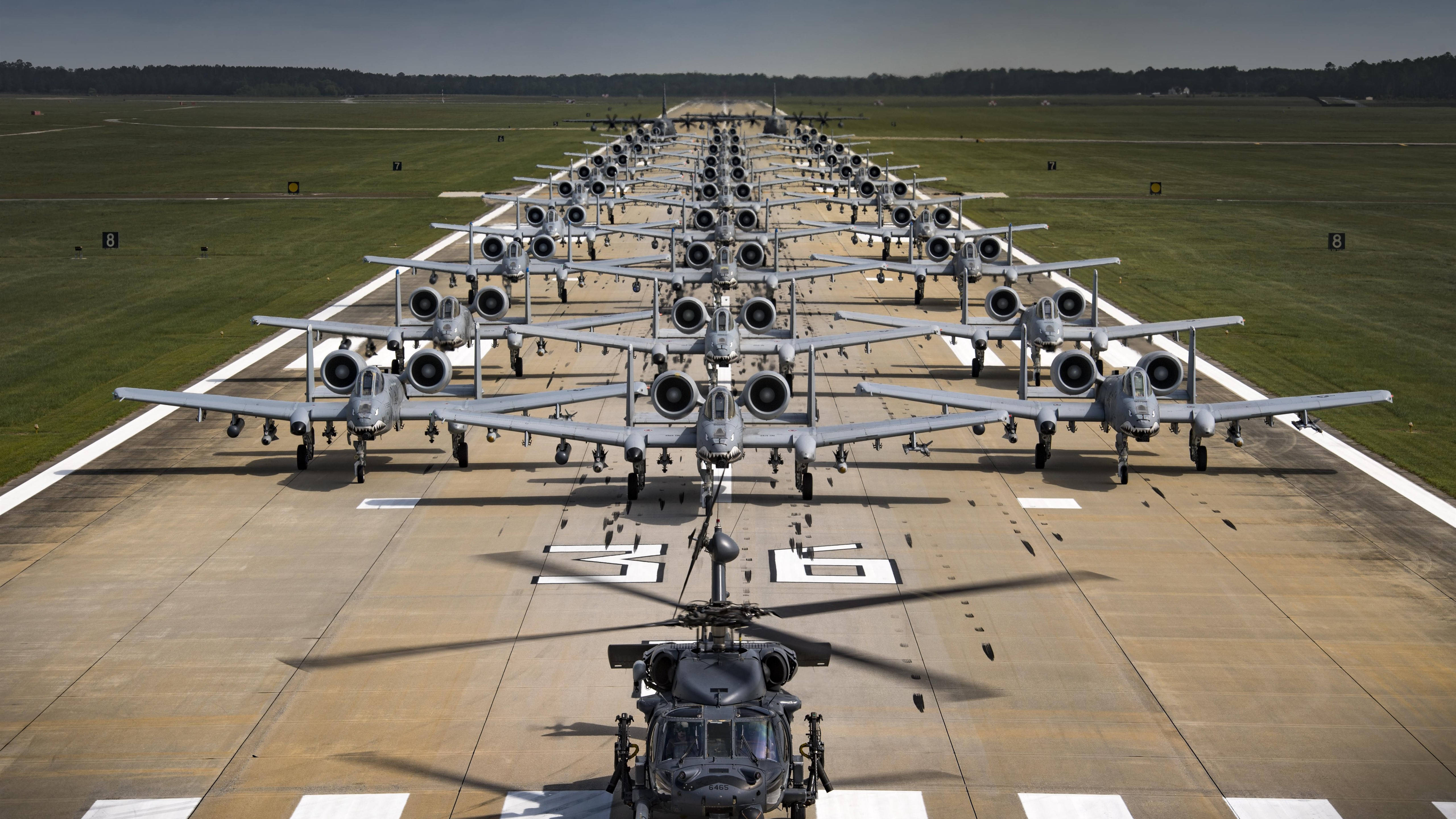 機場上的軍用飛機 壁紙 — 一排排軍用攻擊機排列在停機坪上，前景有一架直升機，天空晴朗。