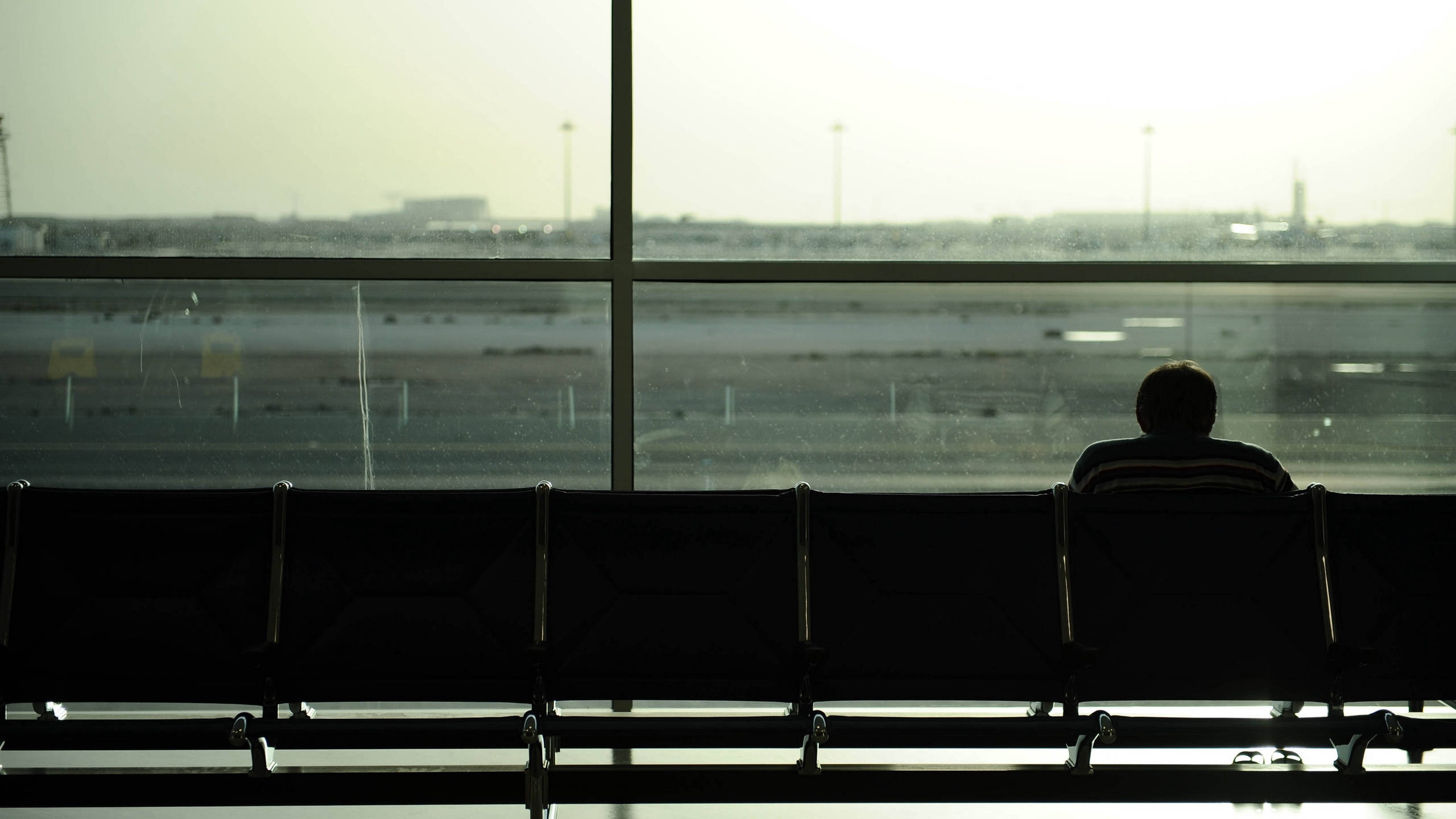 機場休息室內獨坐男子 壁紙 — 一位孤獨的旅客以剪影形式坐在機場長椅上，透過大窗戶凝視著朦朧的停機坪。