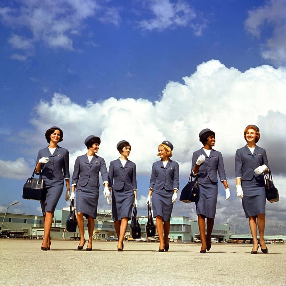 機場停機坪空服員照片 壁紙 — 六名復古航空公司空姐穿著相配的藍色制服，在明亮的藍天和蓬鬆的白雲下自信地走過機場停機坪。