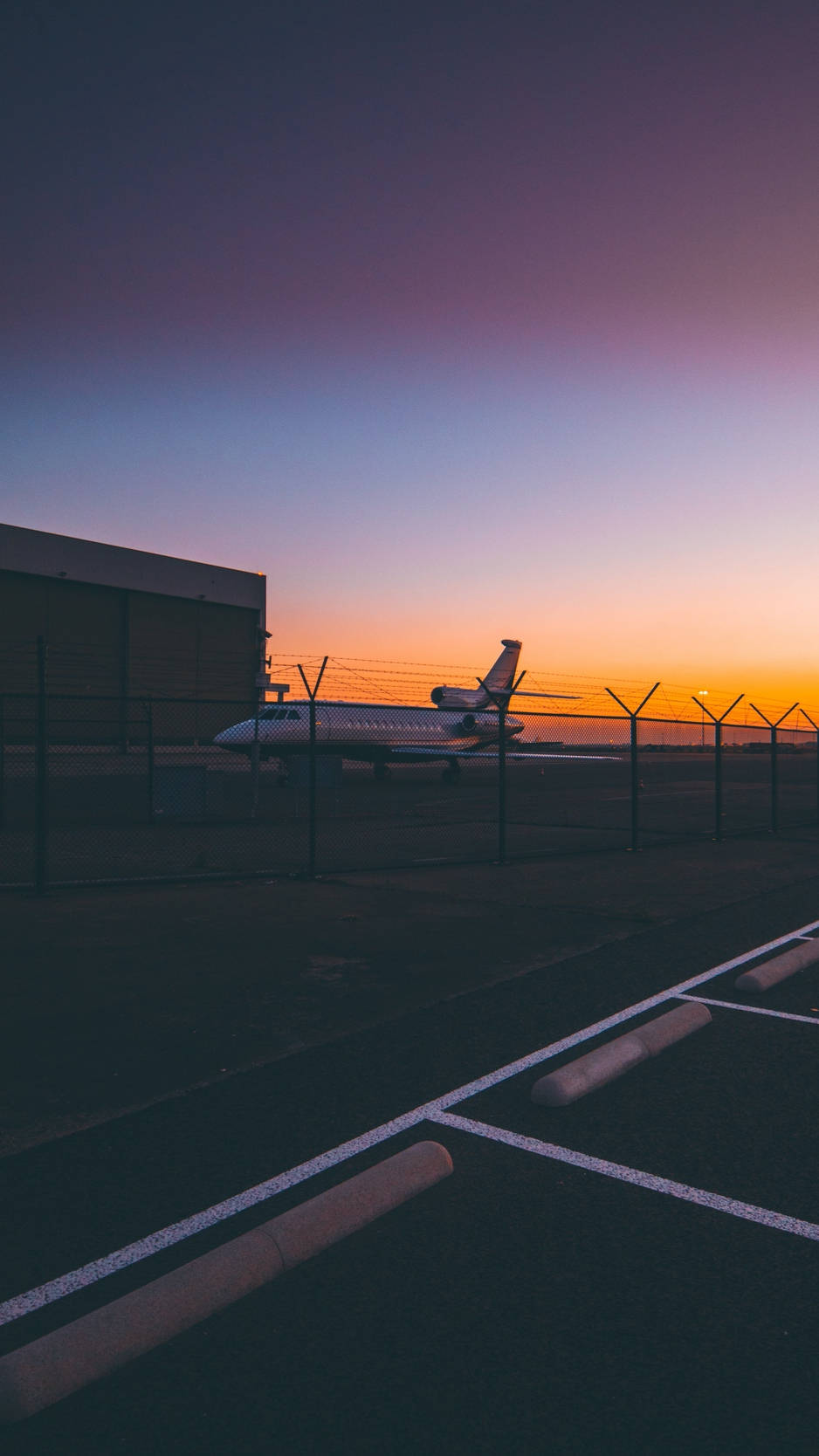 機場日落美景 壁紙 — 一架私人飛機在黃昏時分停泊在圍欄後面，天空呈現紫色、橙色和藍色的漸變色。