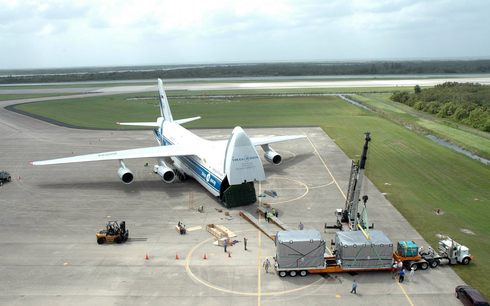 機場貨機與貨車 壁紙 — 一架巨大的白色安東諾夫An-225貨機停在機場停機坪上，地勤人員和重型設備正在裝載巨大的貨物集裝箱。