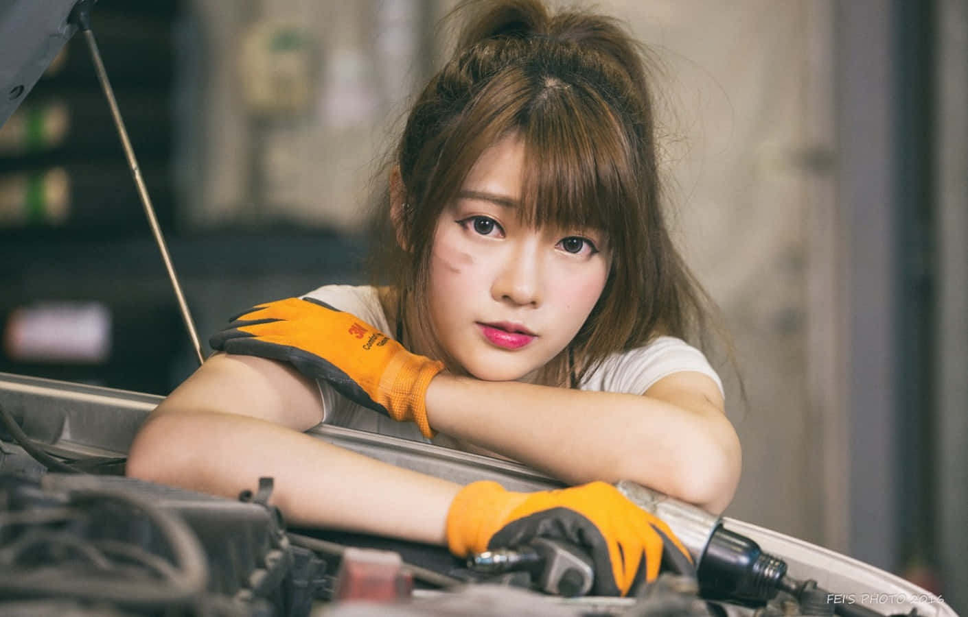 戴著橙色手套的年輕女技師在車庫裡俯身於汽車引擎上。