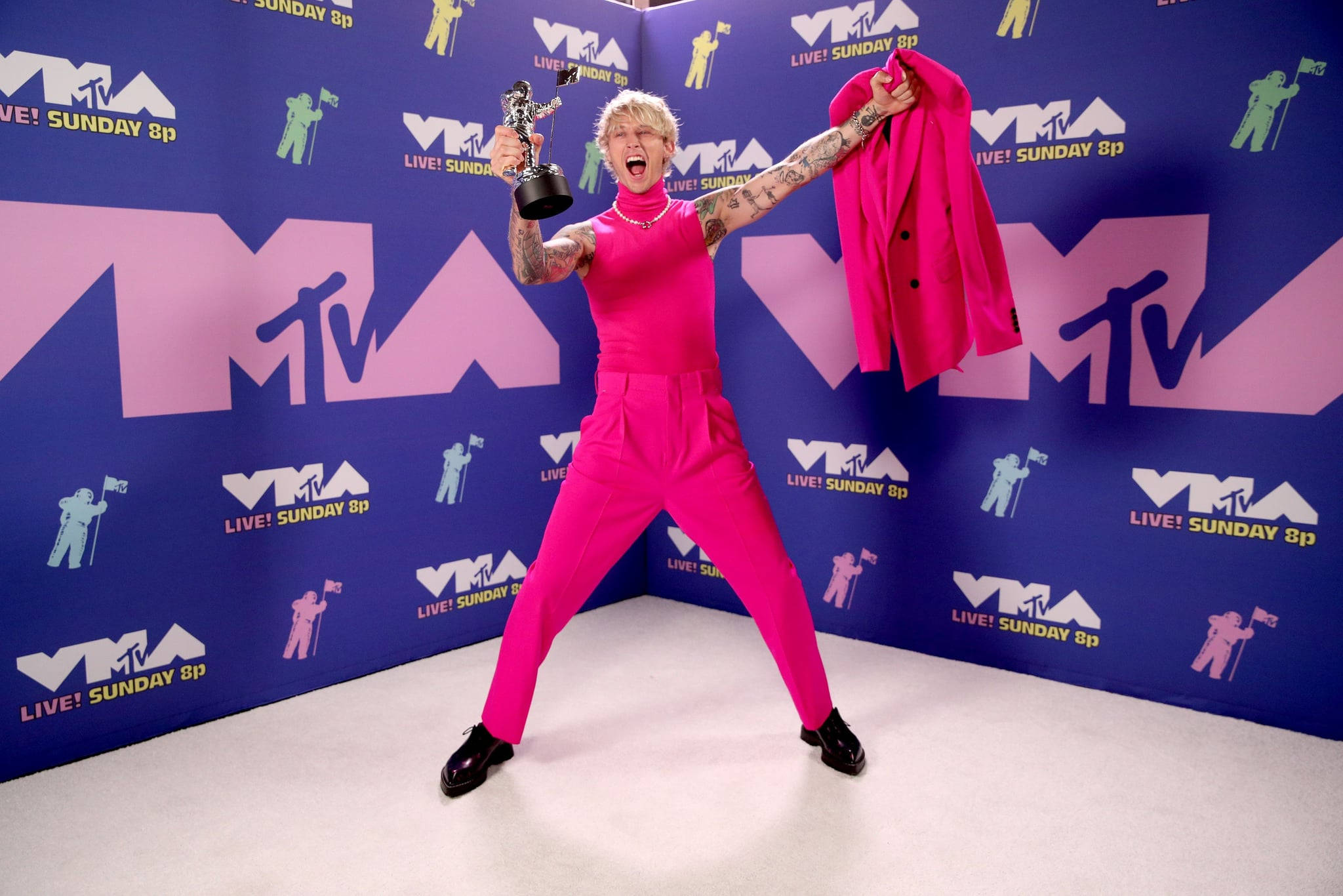 機槍凱爾獲MTV獎項 壁紙 — 一名身穿亮粉紅色連身衣的人，在 MTV 背景下勝利地慶祝，手持獎盃和相配的外套。