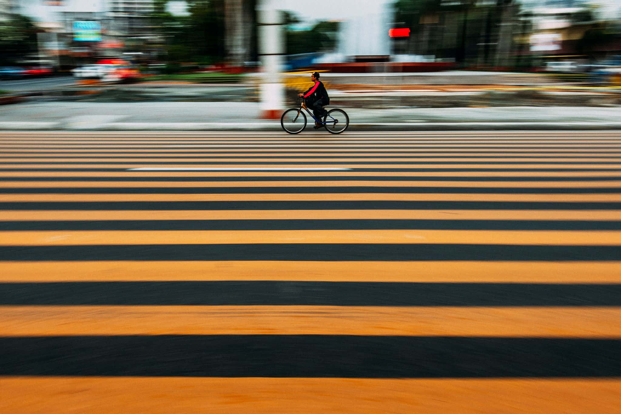 橫向交叉路線圖 壁紙 — 一位正在移動的單車手穿過一條大膽的橙色和黑色條紋行人過路處，背景是模糊的城市景觀。
