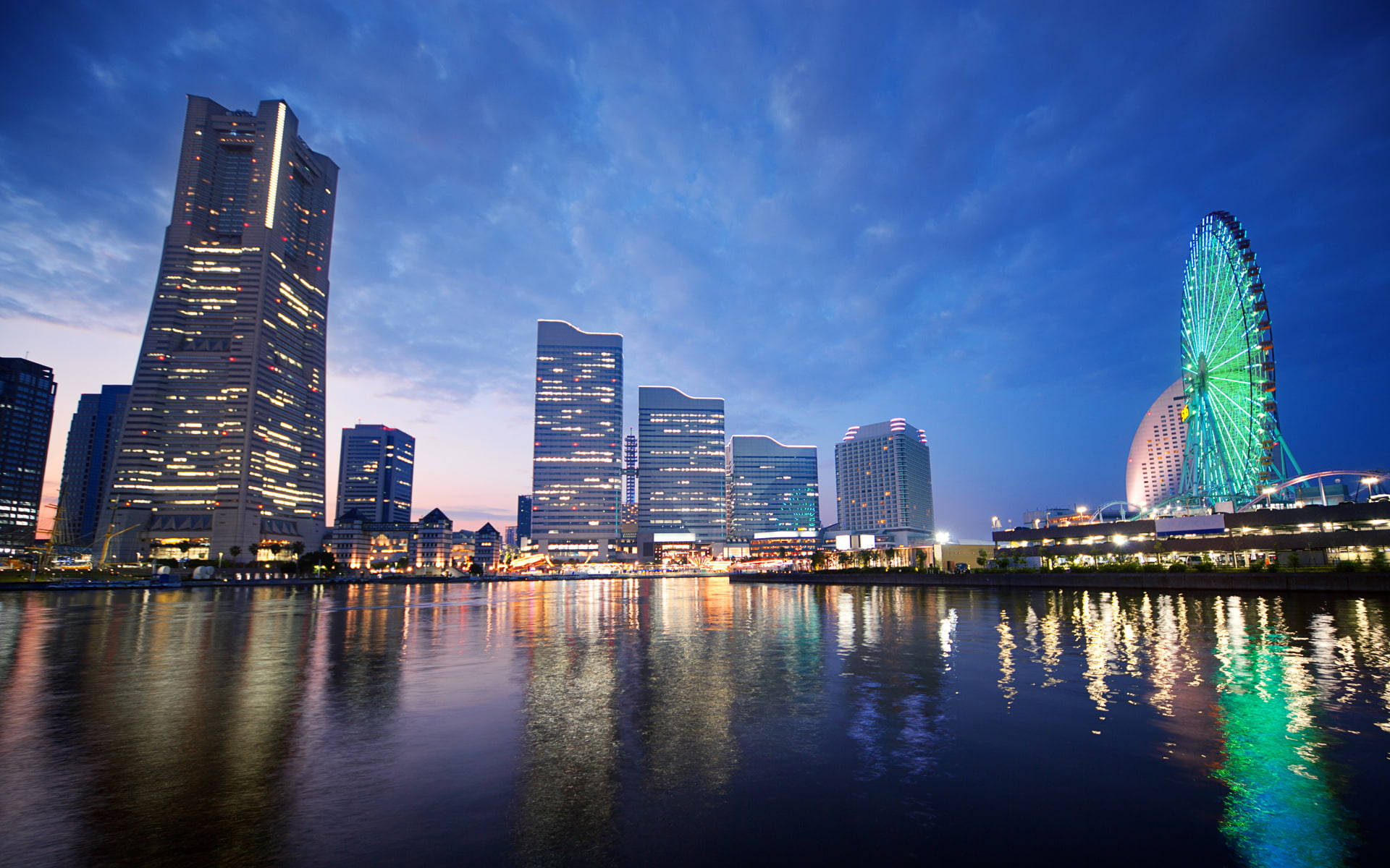 橫濱城市建築與遊樂園黃金時刻 壁紙 — 黃昏時分充滿活力的城市天際線，閃爍的摩天大樓和色彩繽紛的摩天輪倒映在平靜的水面上。