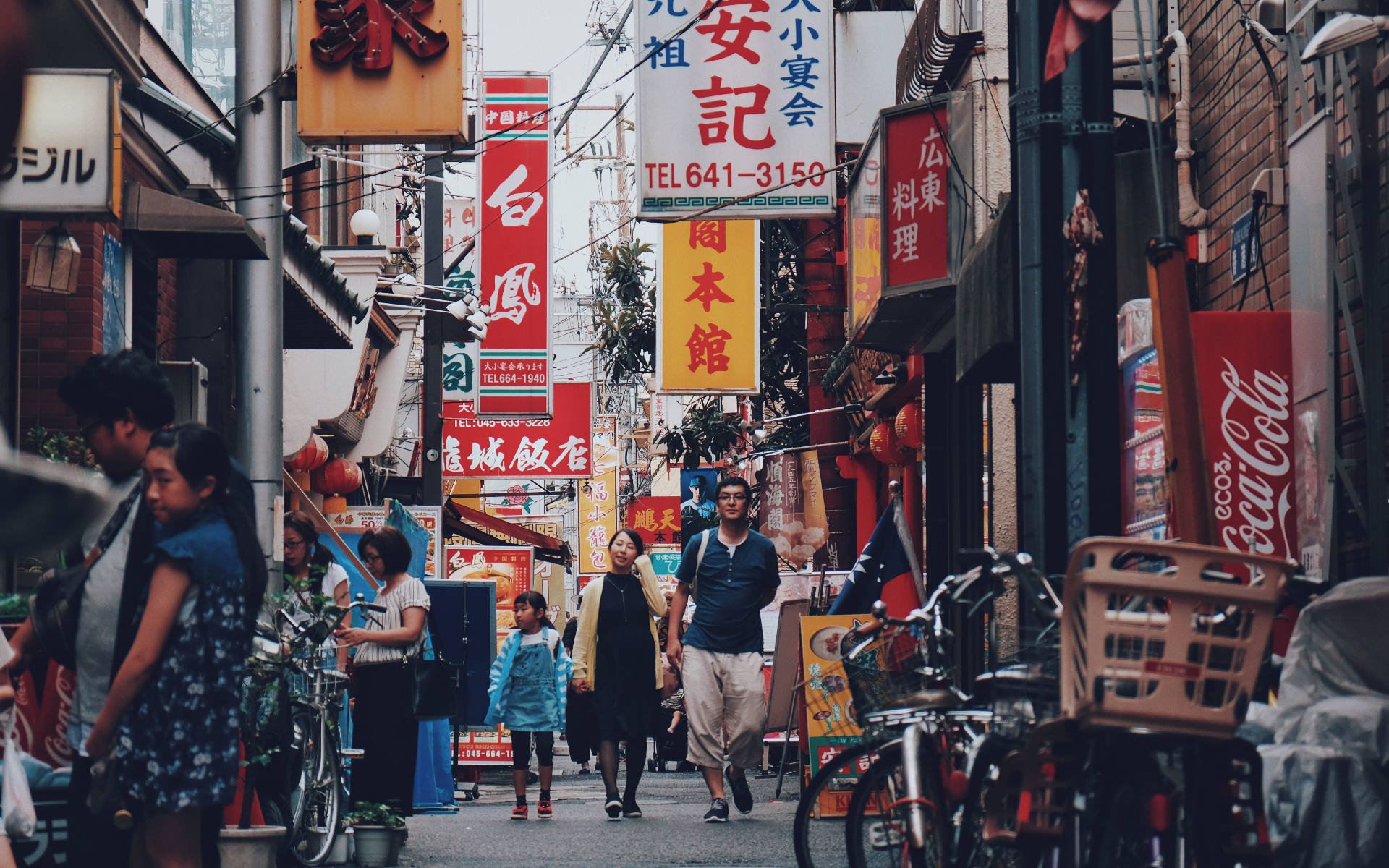 橫濱城市街景 壁紙 — 一條繁忙狹窄的東京小巷，擠滿行人、單車和色彩繽紛的日本招牌，在日光下。