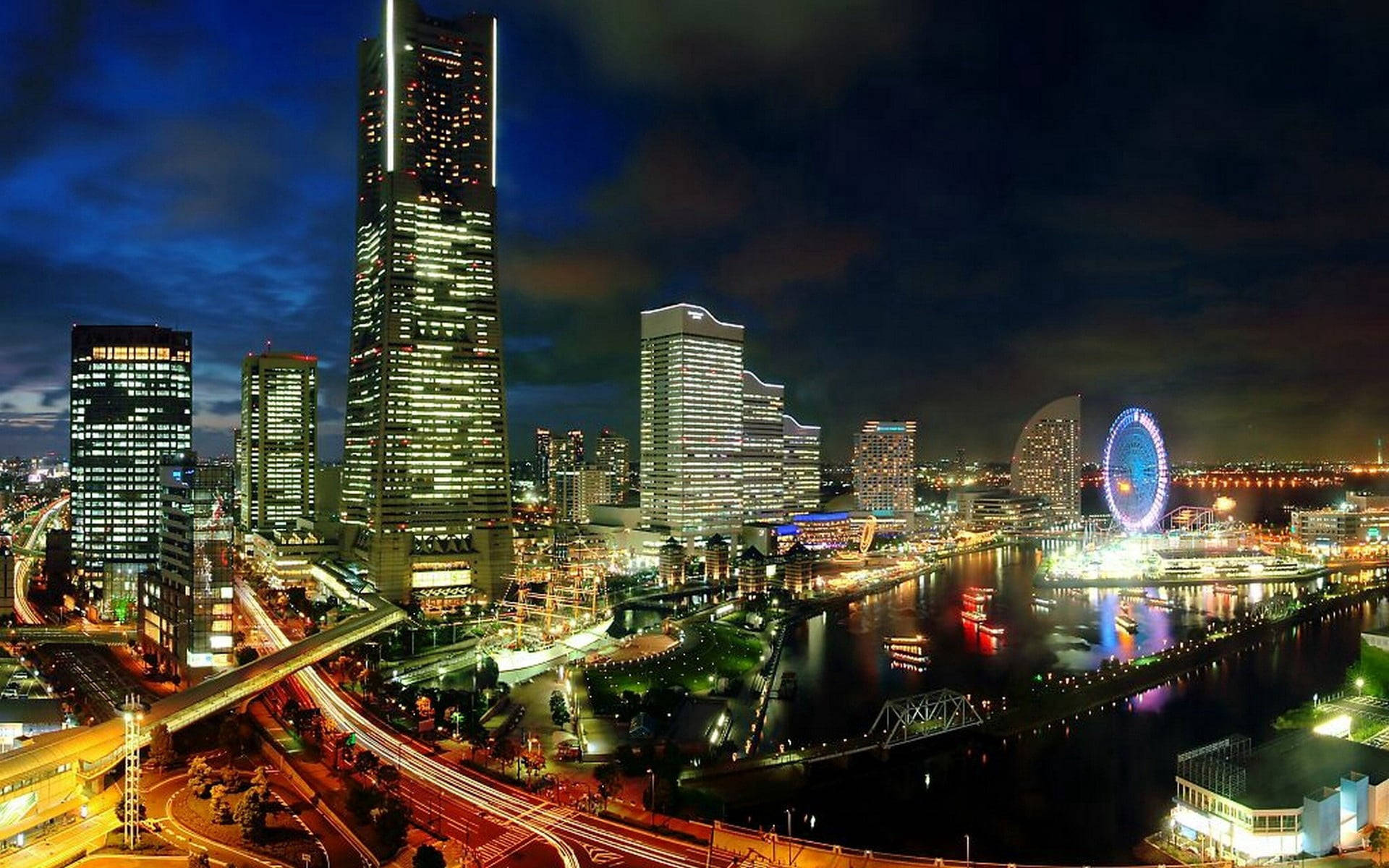 橫濱夜景燈光 壁紙 — 一個令人眼花繚亂的夜間城市景觀，高聳的摩天大樓在黑暗的天空中閃耀，河流反射燈光，一個巨大的摩天輪照亮了海濱。