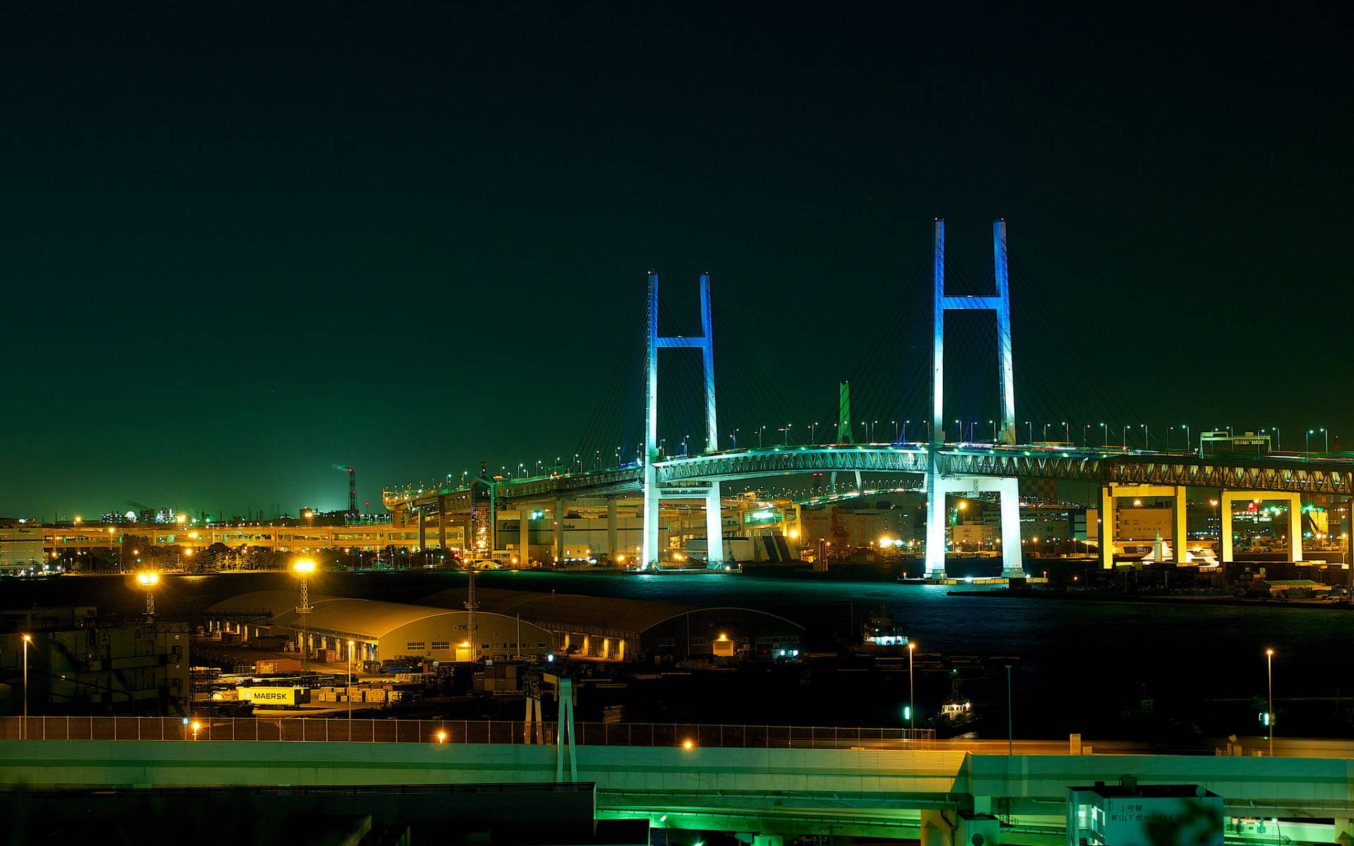 橫濱大橋夜景攝影 壁紙 — 一座高聳的吊橋在繁忙的城市海濱上空，以藍色和綠色的燈光在漆黑的夜空下閃爍。