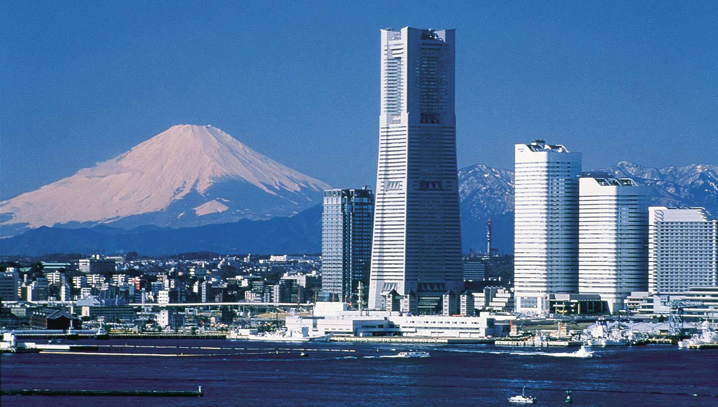 橫濱市與富士山 壁紙 — 白雪覆蓋的富士山在現代東京天際線背後雄偉地升起，玻璃摩天大樓倒映著港口上方的藍天。