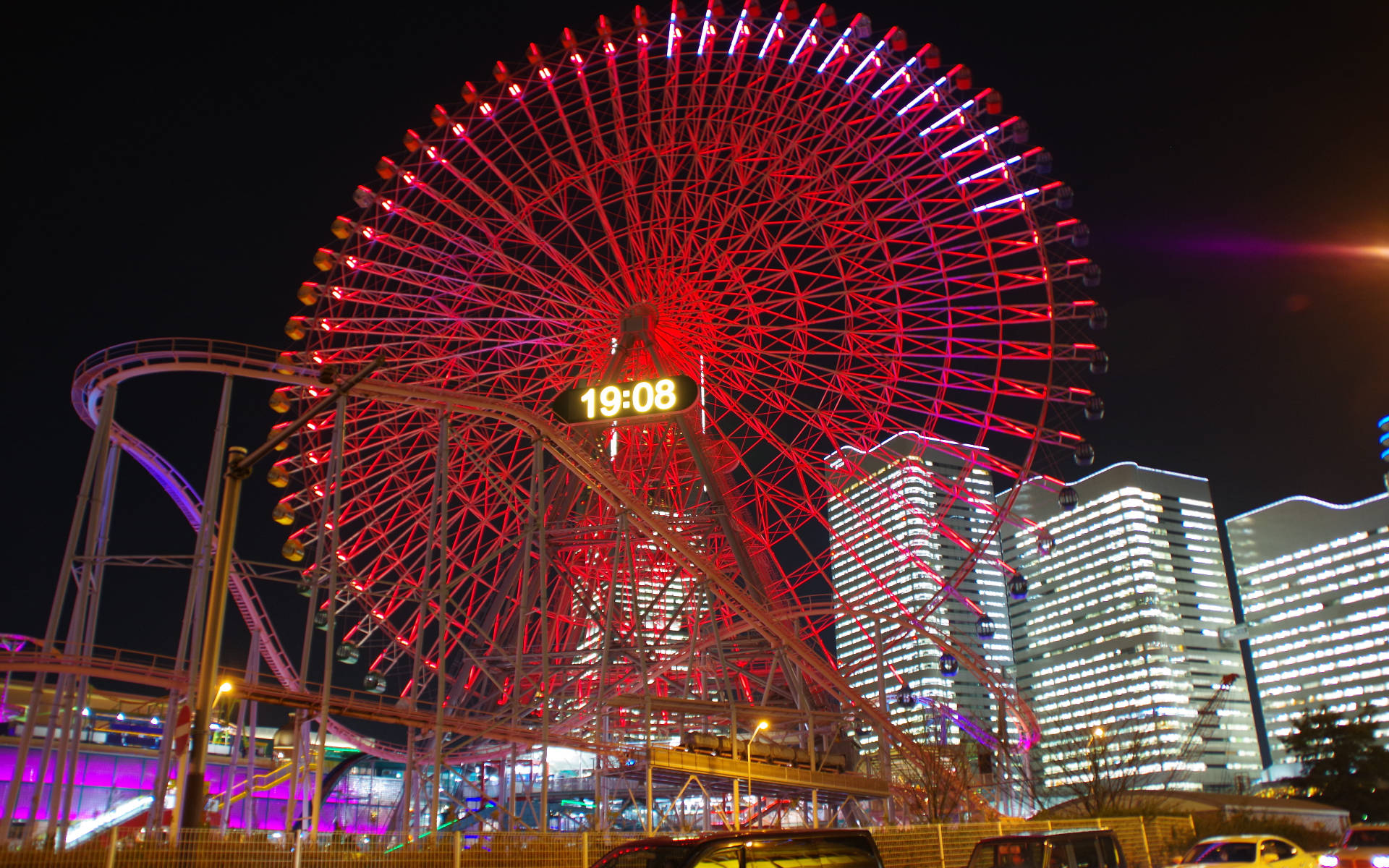 橫濱摩天輪夜景 壁紙 — 一座高聳的紅色摩天輪在黑暗的夜空中閃耀，旁邊是過山車和燈火通明的摩天大樓。