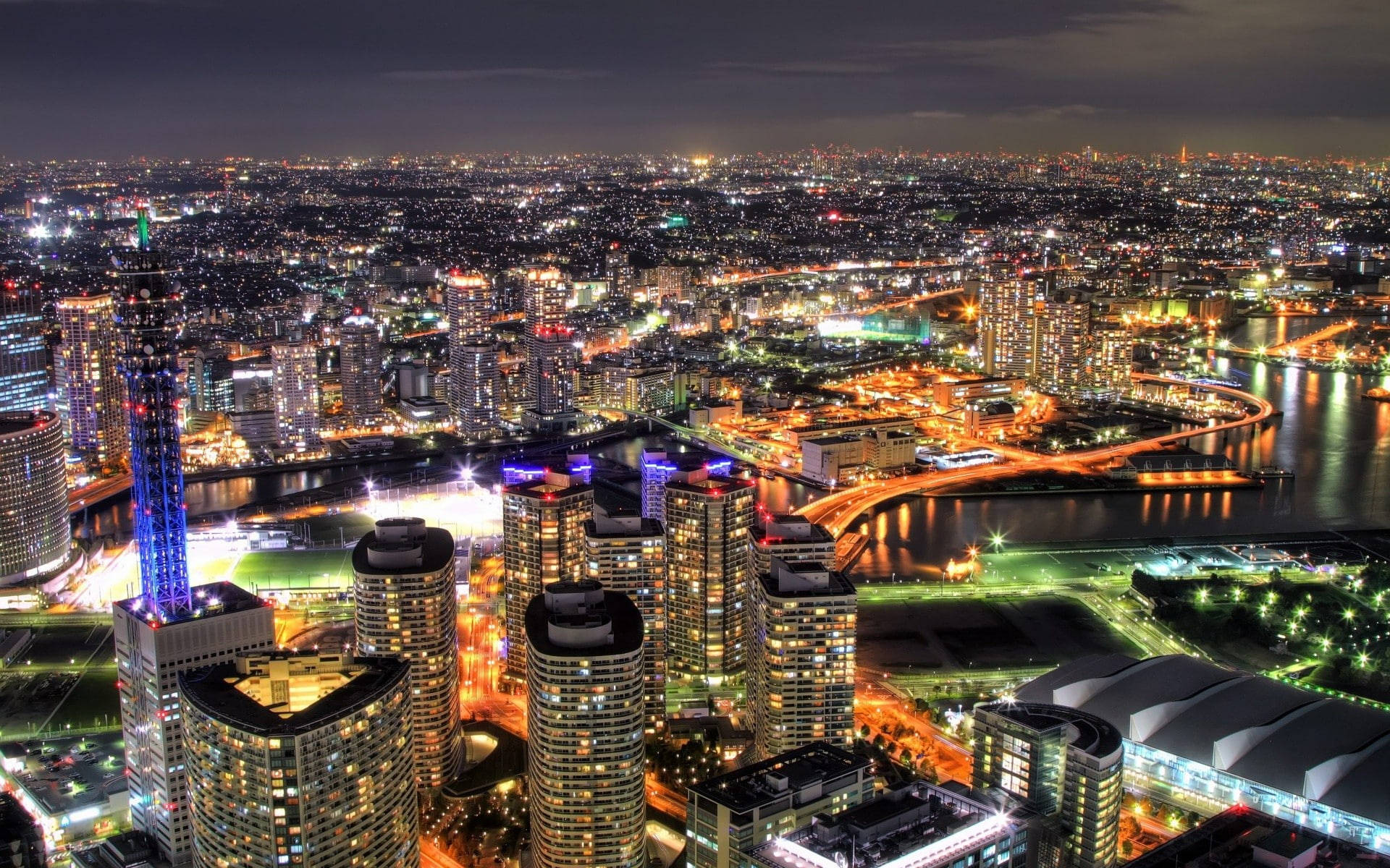 橫濱日本城市天際線 壁紙 — 在黑暗天空下，廣闊城市景觀的夜間航拍，閃爍的摩天大樓、河流和橋樑被照亮。