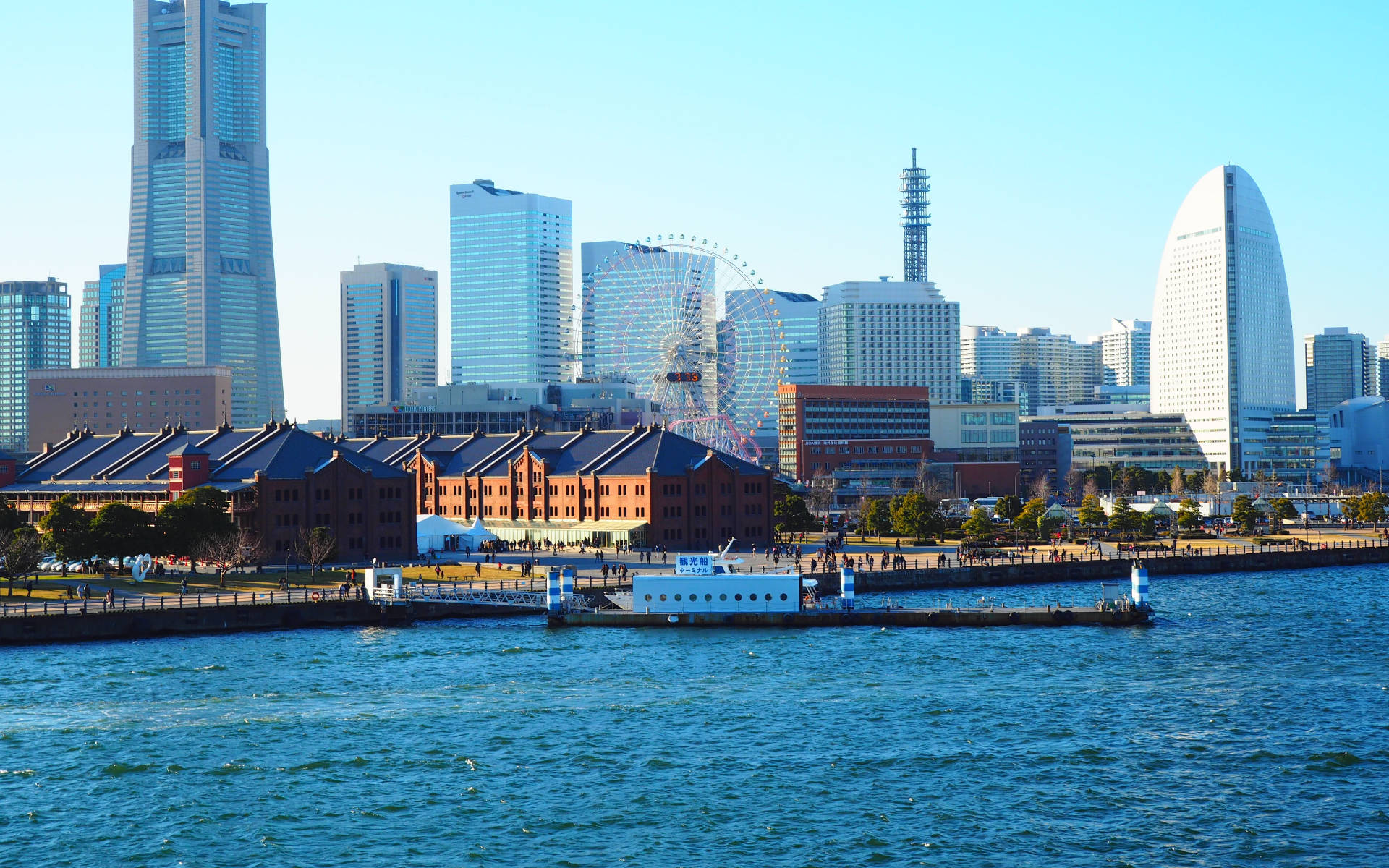 橫濱港口日間 壁紙 — 晴朗日子裡現代城市天際線的景色，高聳的玻璃塔和大型摩天輪背後是歷史悠久的紅磚倉庫，沿著藍色海濱。