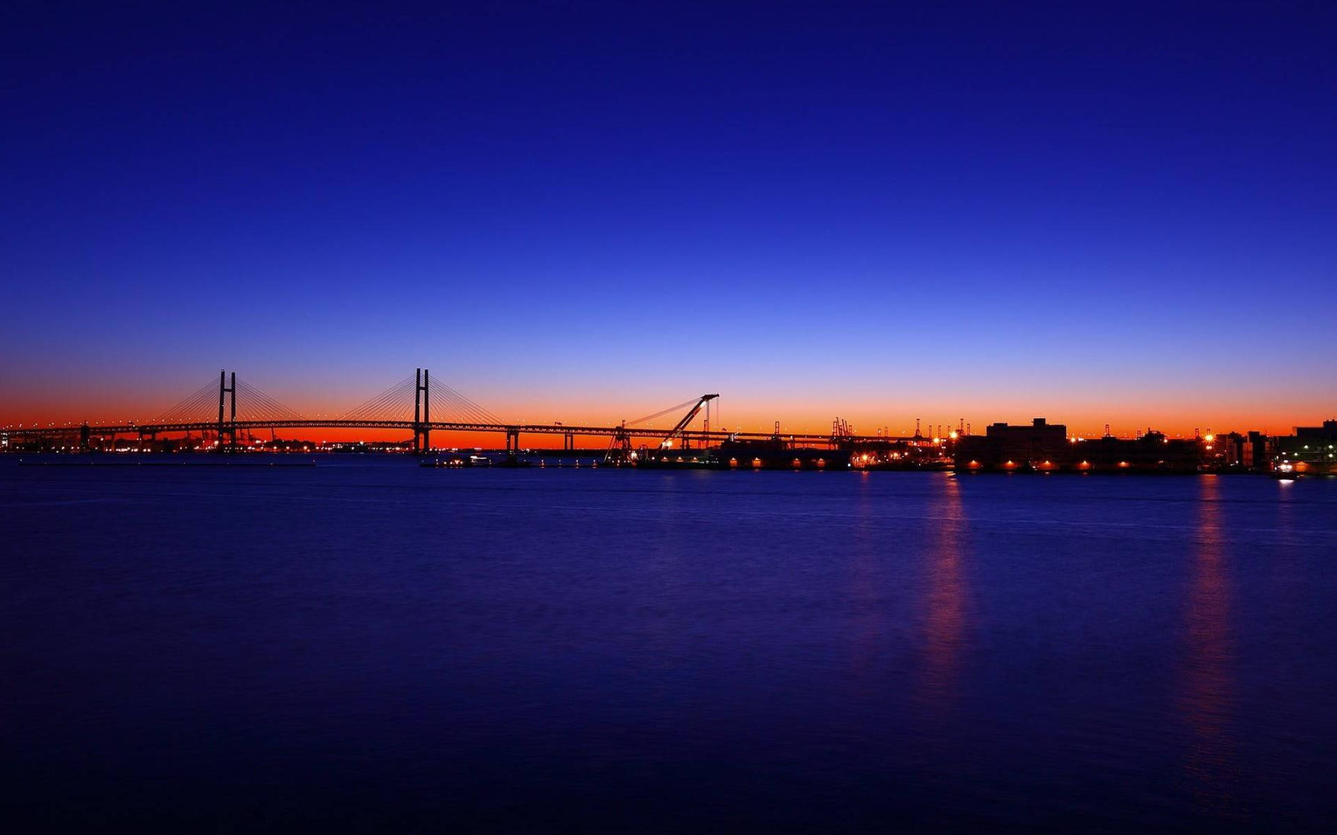 橫濱藍夜城市風光 壁紙 — 一座吊橋在平靜的水面上，以深藍色和火橙色漸變的天空為背景，呈現出令人驚嘆的黃昏景色。