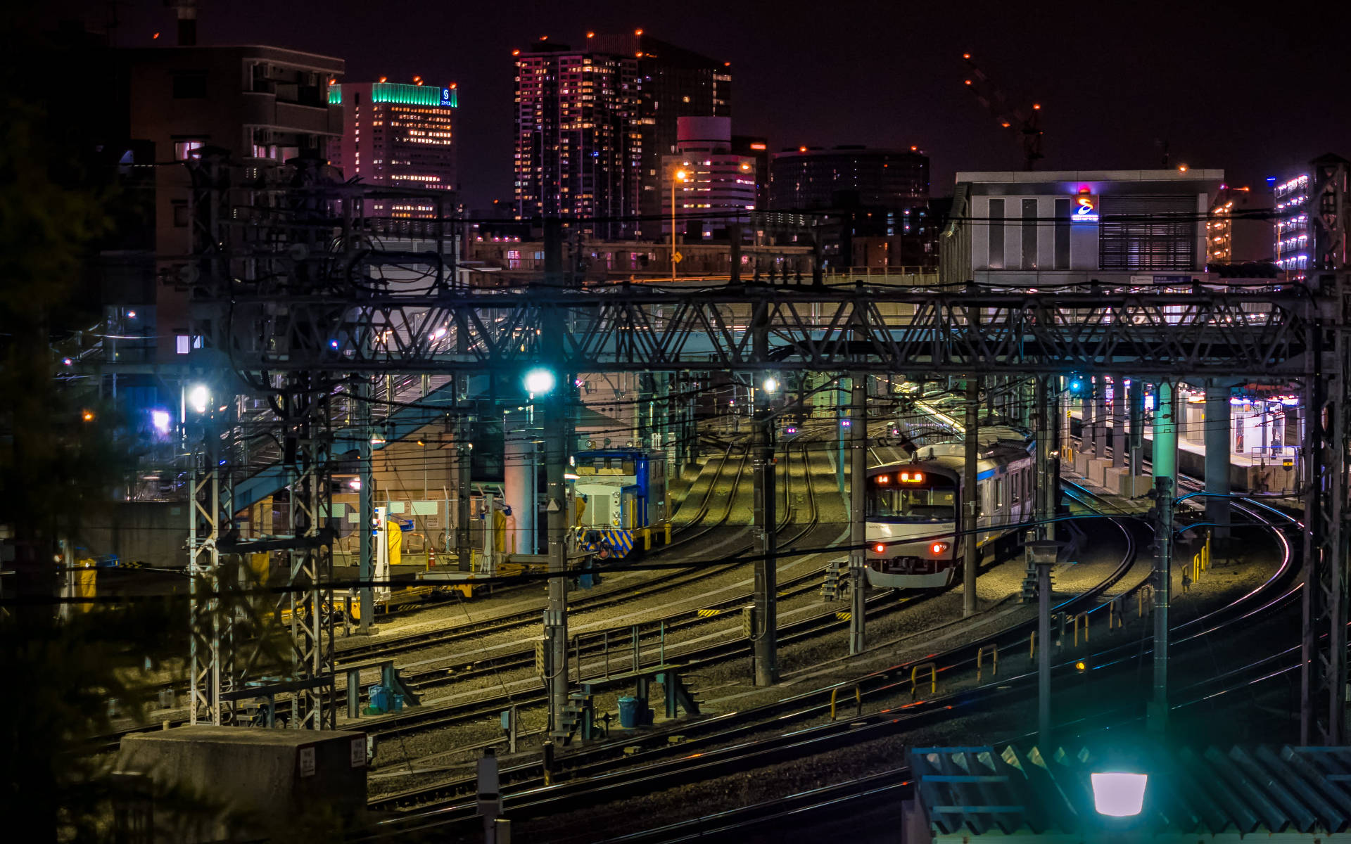 橫濱鐵路夜景 壁紙 — 火車在夜間穿過霓虹燈閃爍的城市天際線下複雜的軌道網絡。