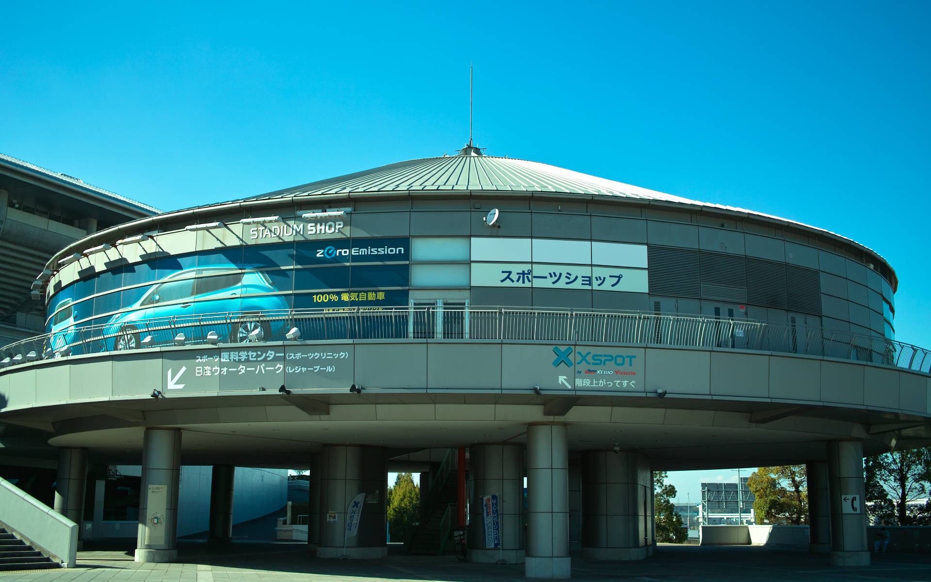 橫濱體育場 壁紙 — 一座現代圓形體育館建築，玻璃幕牆，藍色汽車廣告，在清澈藍天下。