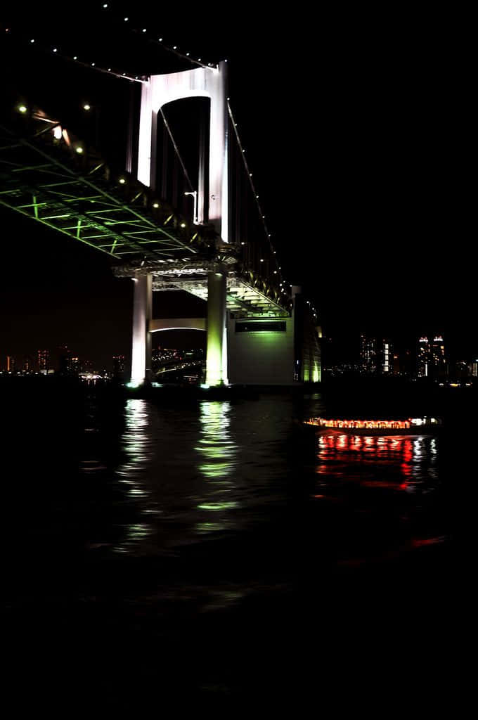 橫跨山谷的魔法彩虹橋 壁紙 — 一座高聳的照明橋樑在夜間倒映在黑暗的水面上，附近有一艘發光的船。