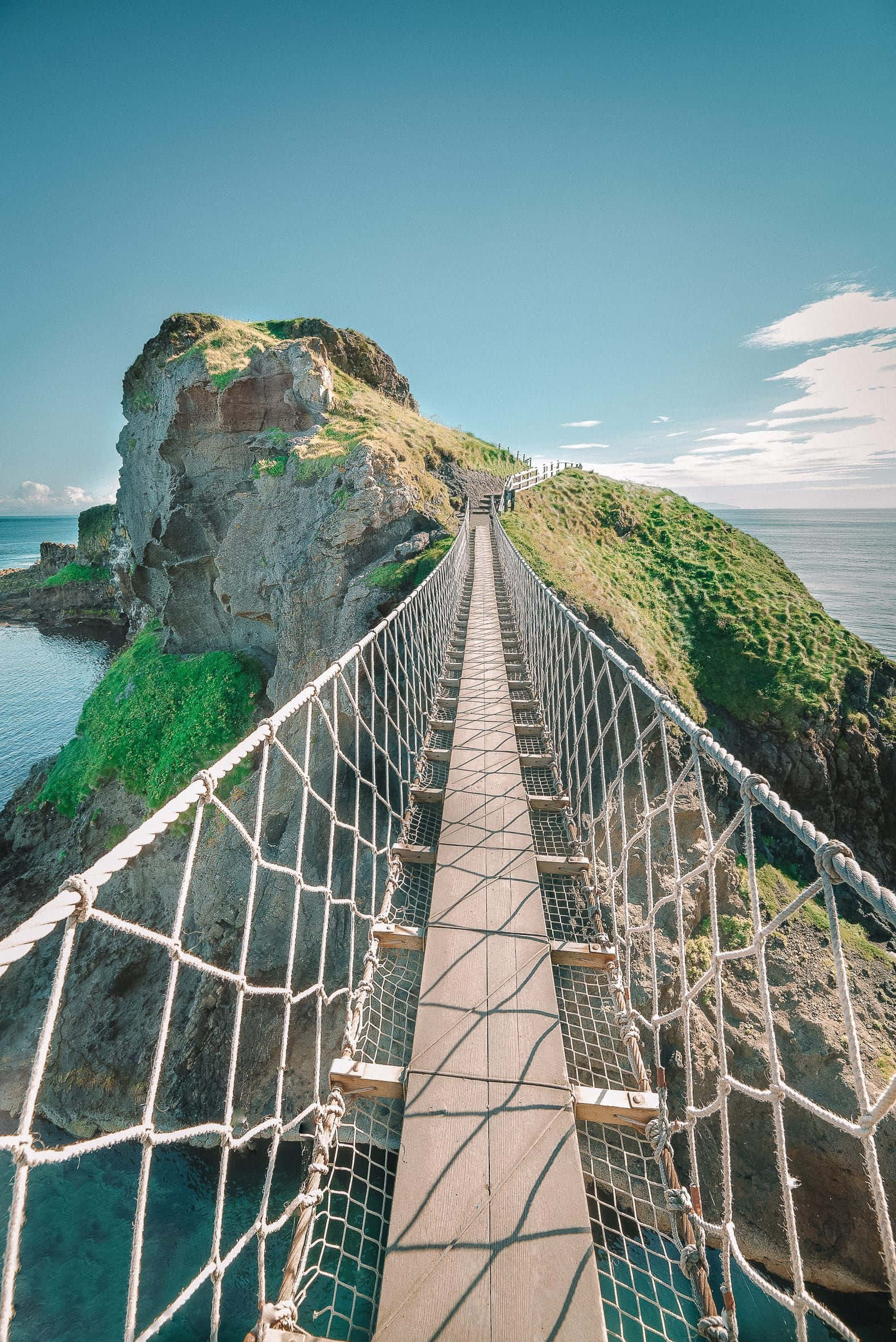 橫跨飛瀑的木橋風景 壁紙 — 一條繩索吊橋橫跨碧綠色的海水，連接到一片草地懸崖，上方是清澈的藍天。
