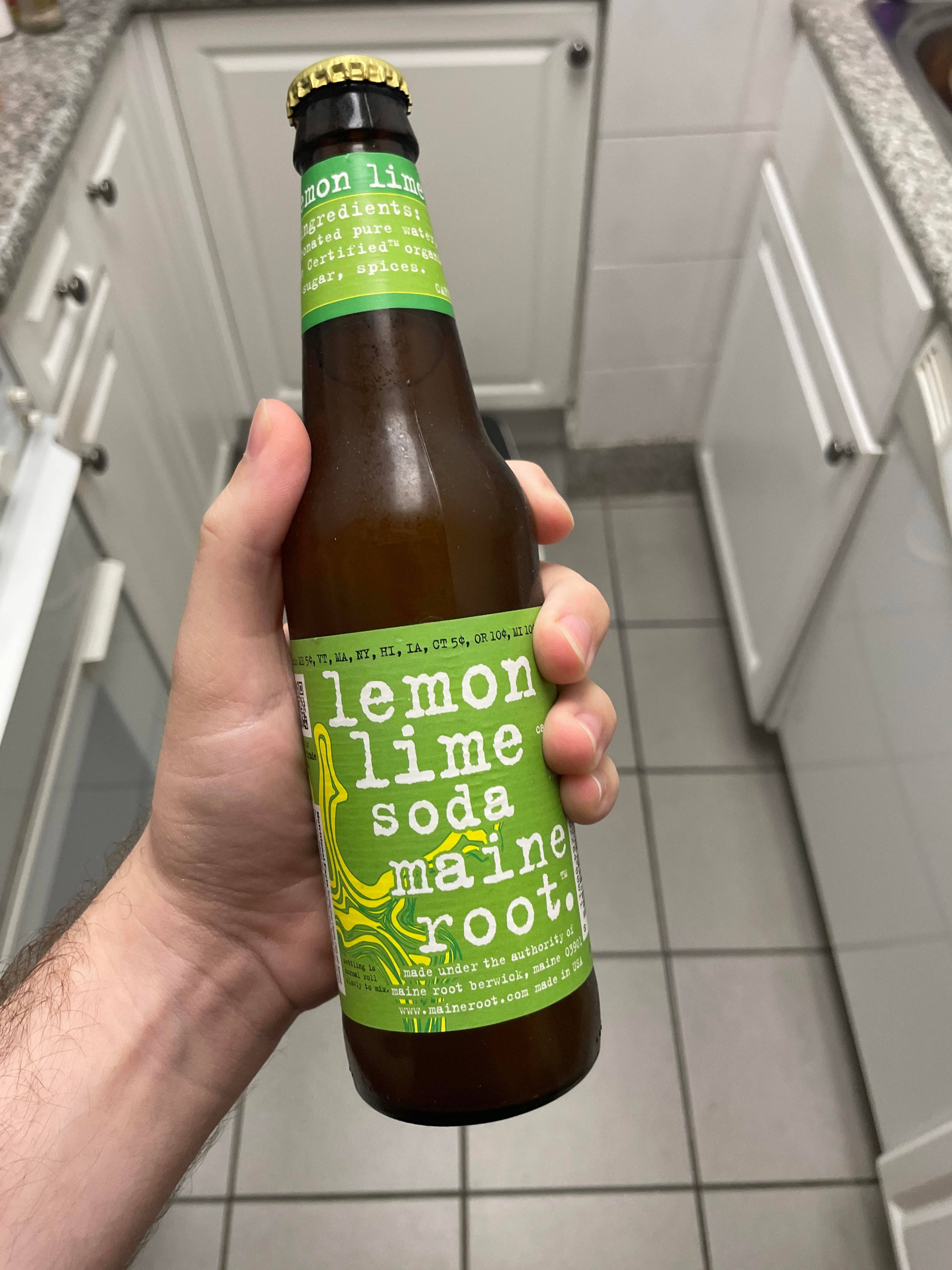 檸檬青檸味蘇打緬因州根汁飲品 壁紙 — 一隻手在明亮的廚房裡拿著一個綠色標籤的Maine Root檸檬青檸汽水瓶，廚房有白色櫥櫃和瓷磚地板。