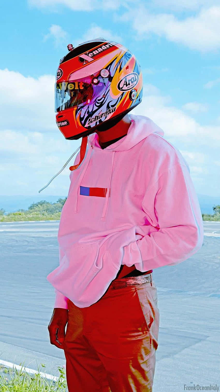 欣賞 Frank Ocean《Blonde》專輯的藝術美感 壁紙 — 一個人穿著鮮豔的粉紅色連帽衫和紅色褲子，在明亮的藍天下戴著色彩繽紛的賽車頭盔站在賽車場上。
