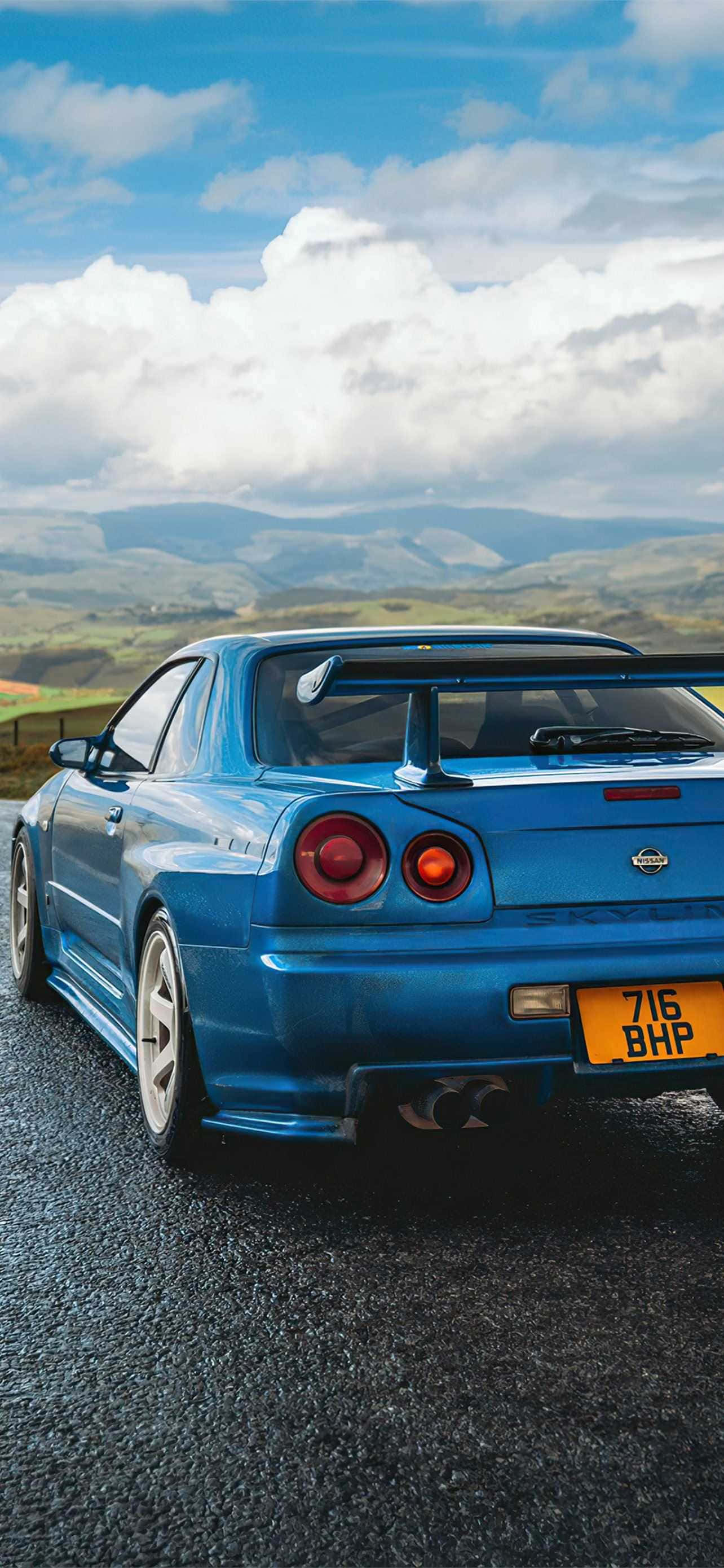欣賞 Nissan Skyline R33 GTR 的流線美學 壁紙 — 一架充滿活力的藍色日產Skyline R34 GT-R停泊在碎石路上，背景是起伏的綠色山丘和蓬鬆的白雲，天空湛藍明亮。