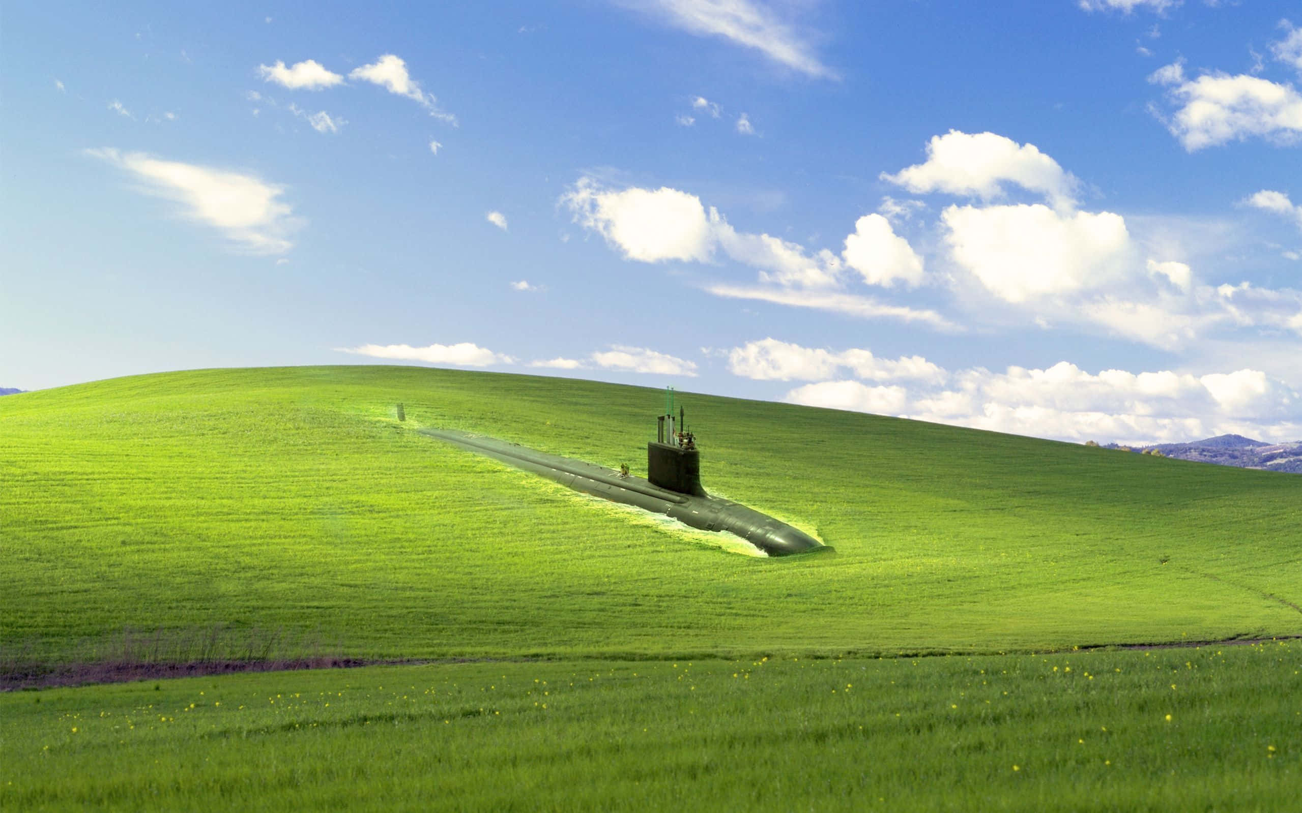 欣賞 Windows XP 的美感 壁紙 — 一艘深色潛艇在充滿活力的綠色山丘上休息，上方是明亮的藍天和蓬鬆的白雲。