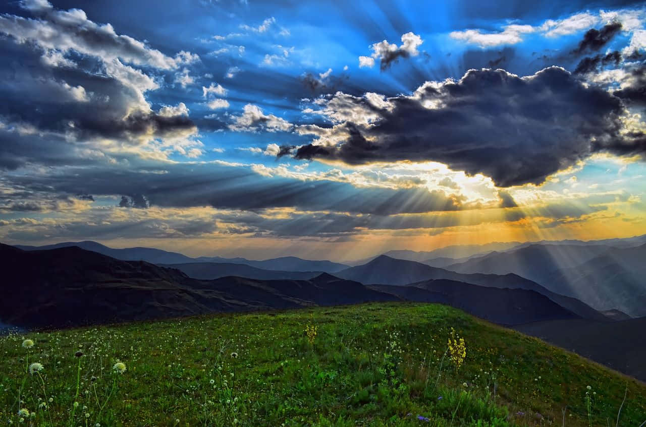 欣賞初春早晨的自然美景 壁紙 — 陽光穿過戲劇性的雲層，照耀在連綿起伏的山脈上，前景是一片充滿活力的綠色草地。