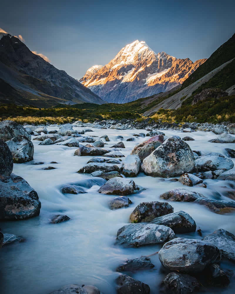 一條河流過岩石，白雪覆蓋的山峰在晨光中閃耀。