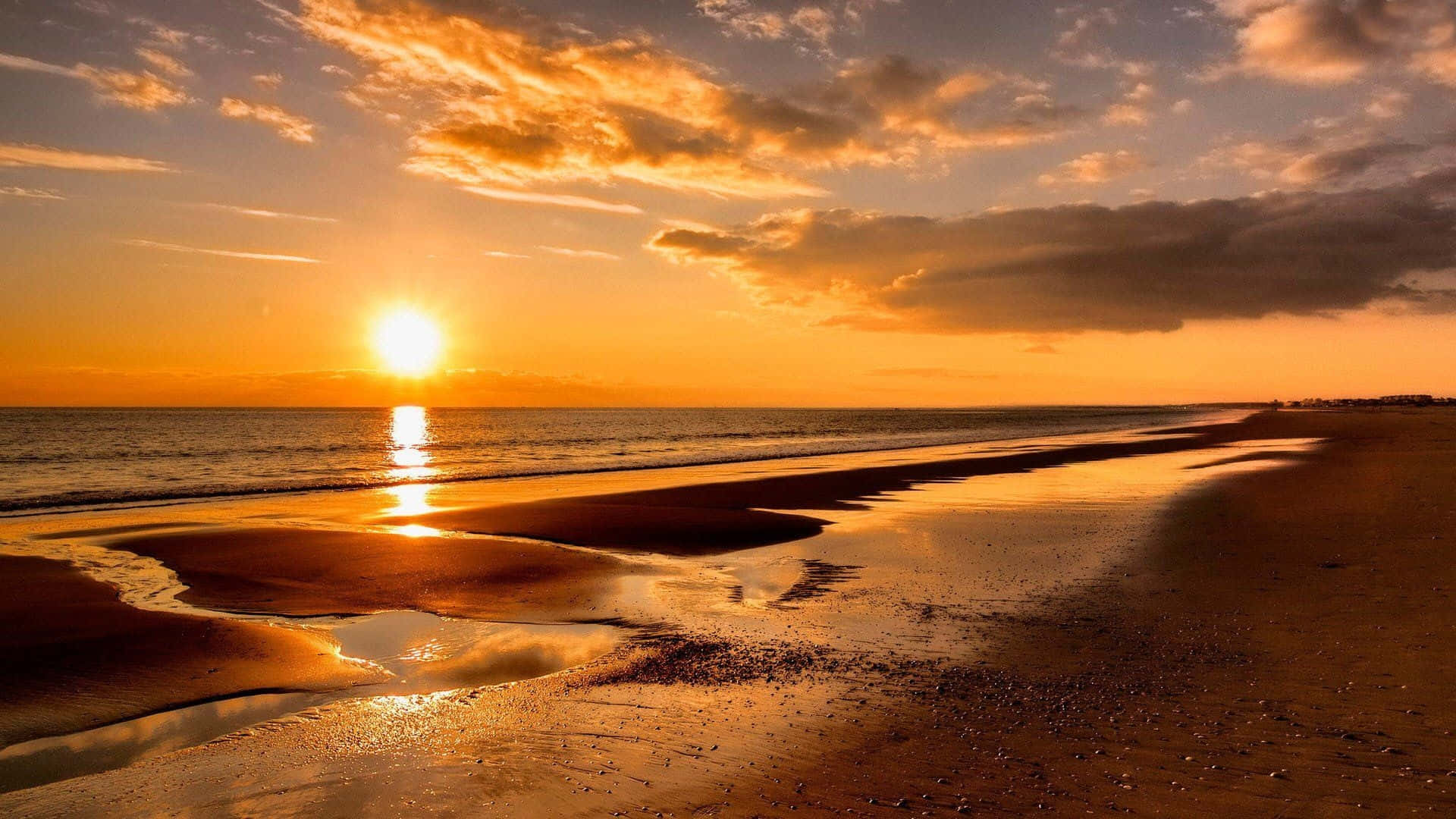 欣賞壯觀海灘日落之美 壁紙 — 金色夕陽照耀在海浪上，沖刷著濕潤的沙灘，天空有散落的雲朵。