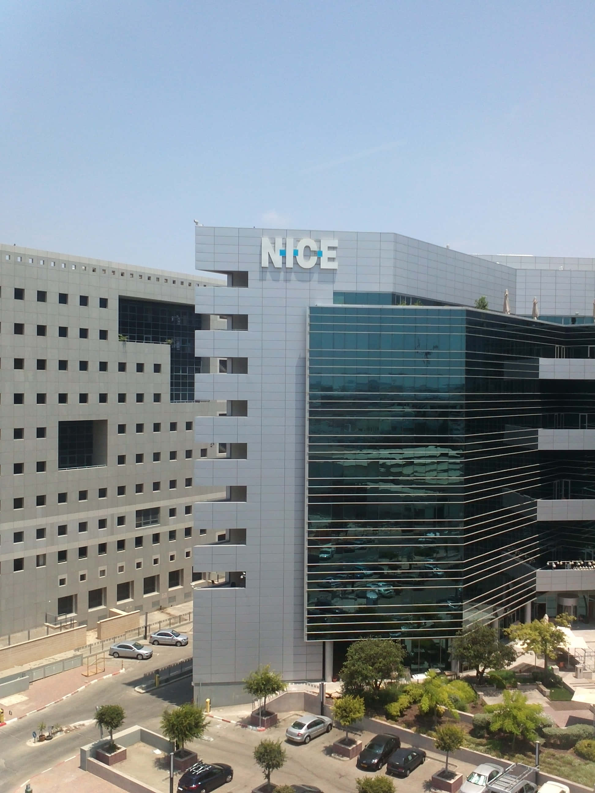 欣賞尼斯全景 壁紙 — 現代玻璃鋼筋企業大樓，帶有「NICE」標誌，在晴朗藍天下，下方有停泊的汽車和樹木。