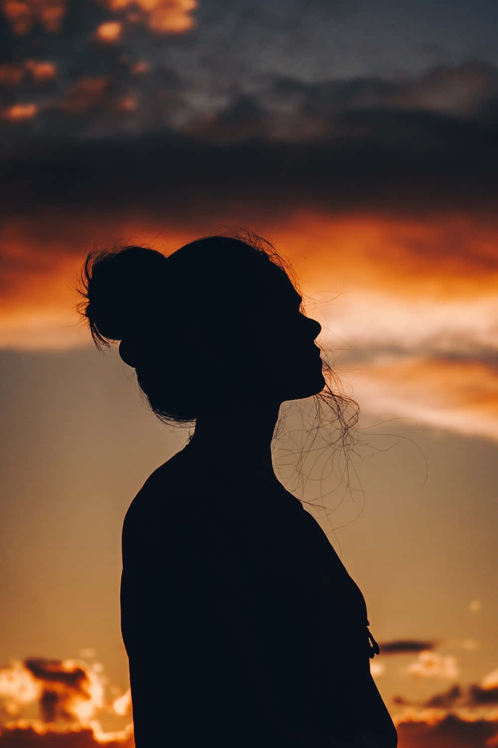 欣賞日落時分 壁紙 — 一個頭髮盤成髮髻的女人的剪影，在熾熱的橙色和紫色日落天空下向上凝望。