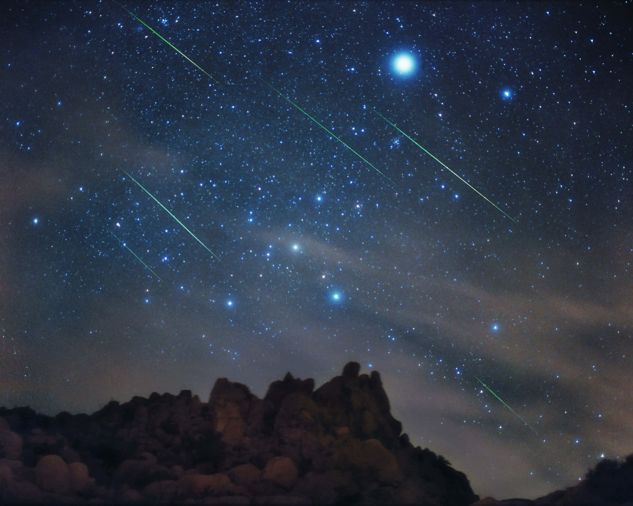 欣賞流星照亮夜空 壁紙 — 星光燦爛的夜空，明亮的流星在崎嶇岩石地形上劃過，下方是一輪明亮的月亮。