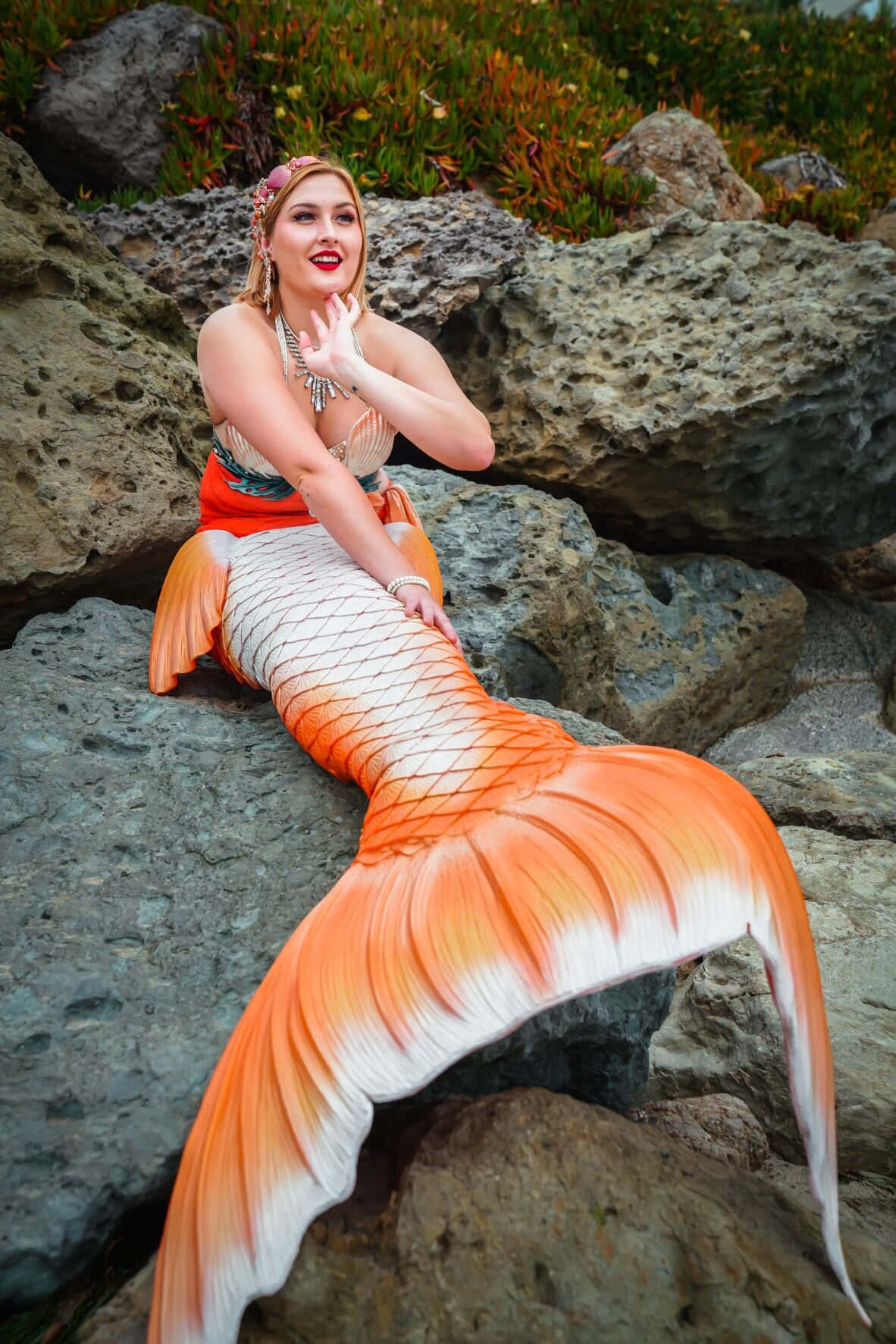 一位穿著橙白色美人魚尾巴的女性坐在岩石上。