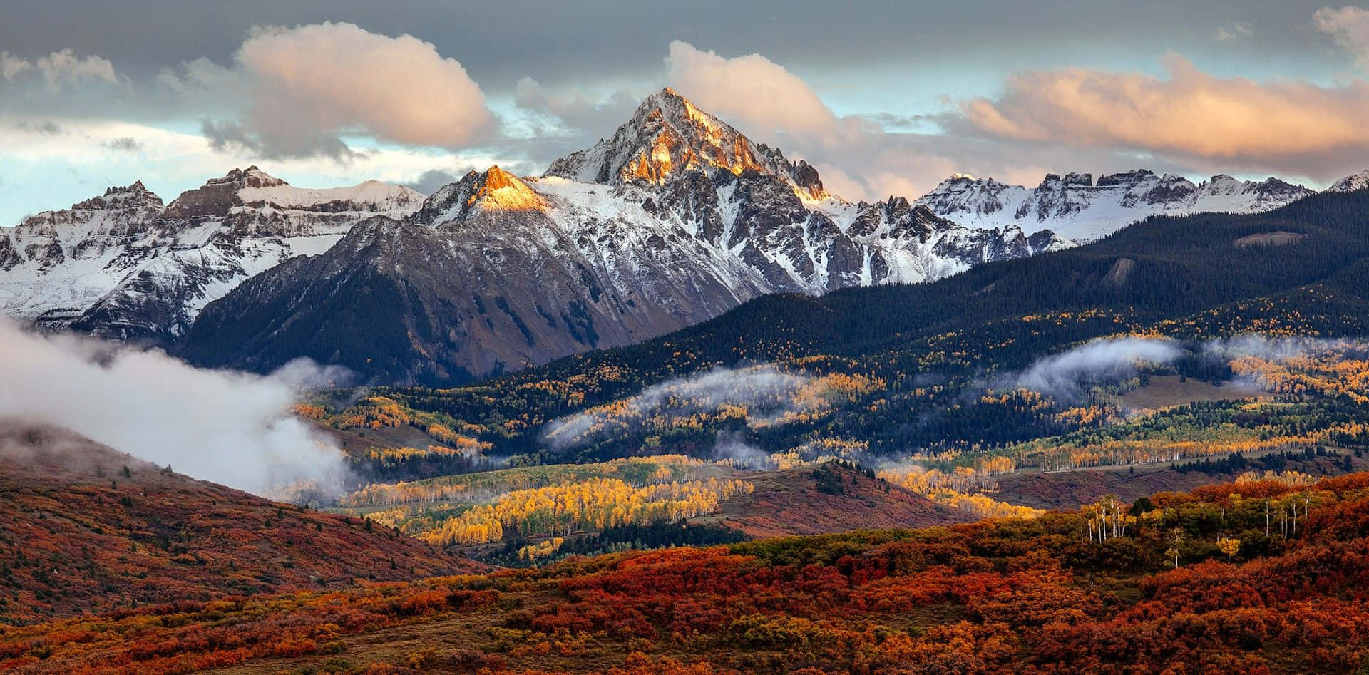 欣賞科羅拉多山的壯麗美景 壁紙 — 白雪覆蓋的山峰在日落時閃耀著金色光芒，下方是秋日紅黃交織、如火燃燒的森林。