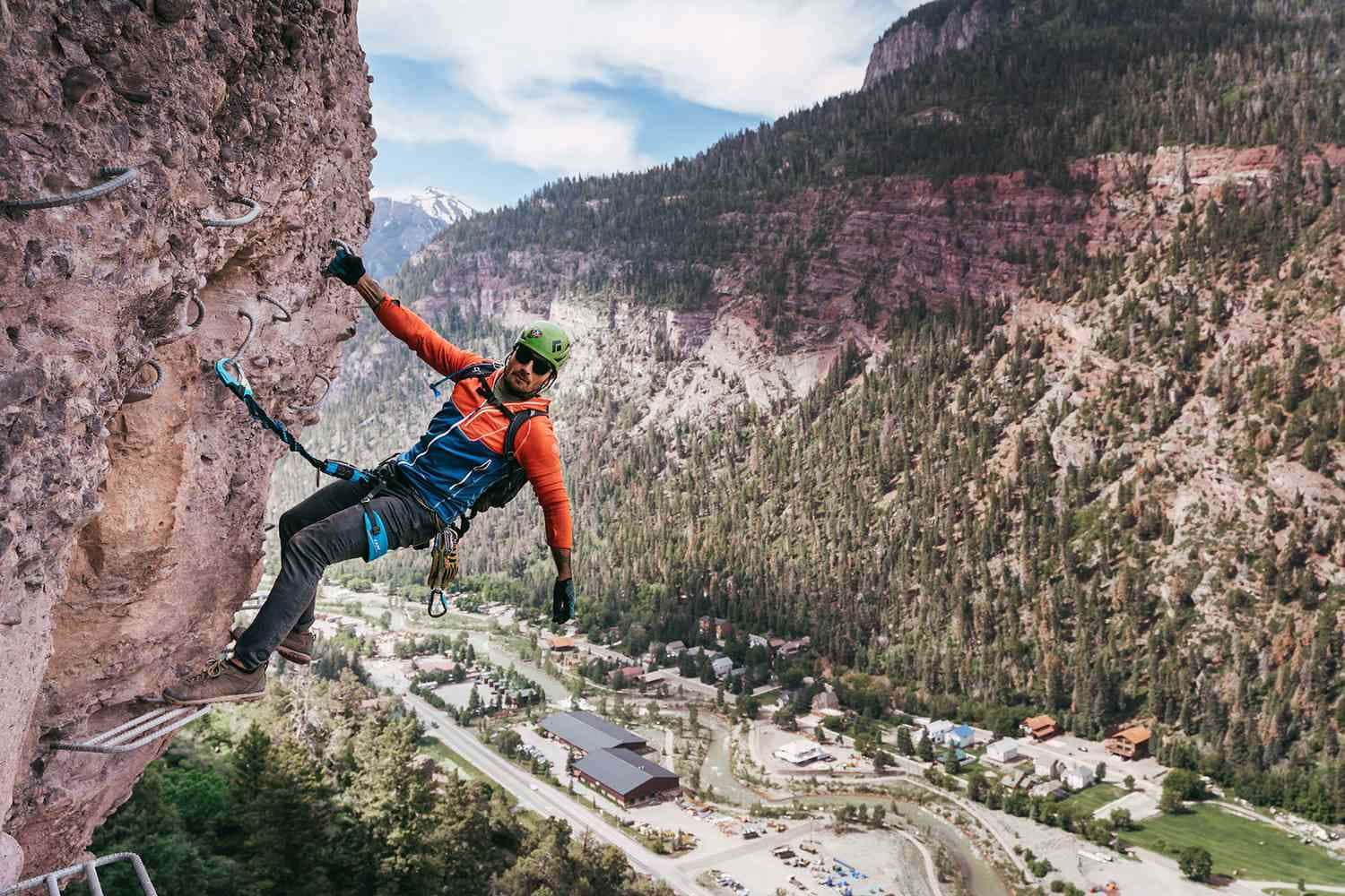 欣賞科羅拉多山的美景 壁紙 — 一名穿著橙色裝備的攀岩者正在攀登陡峭的懸崖，俯瞰著森林覆蓋的山谷和遠處白雪覆蓋的山峰，天空湛藍。