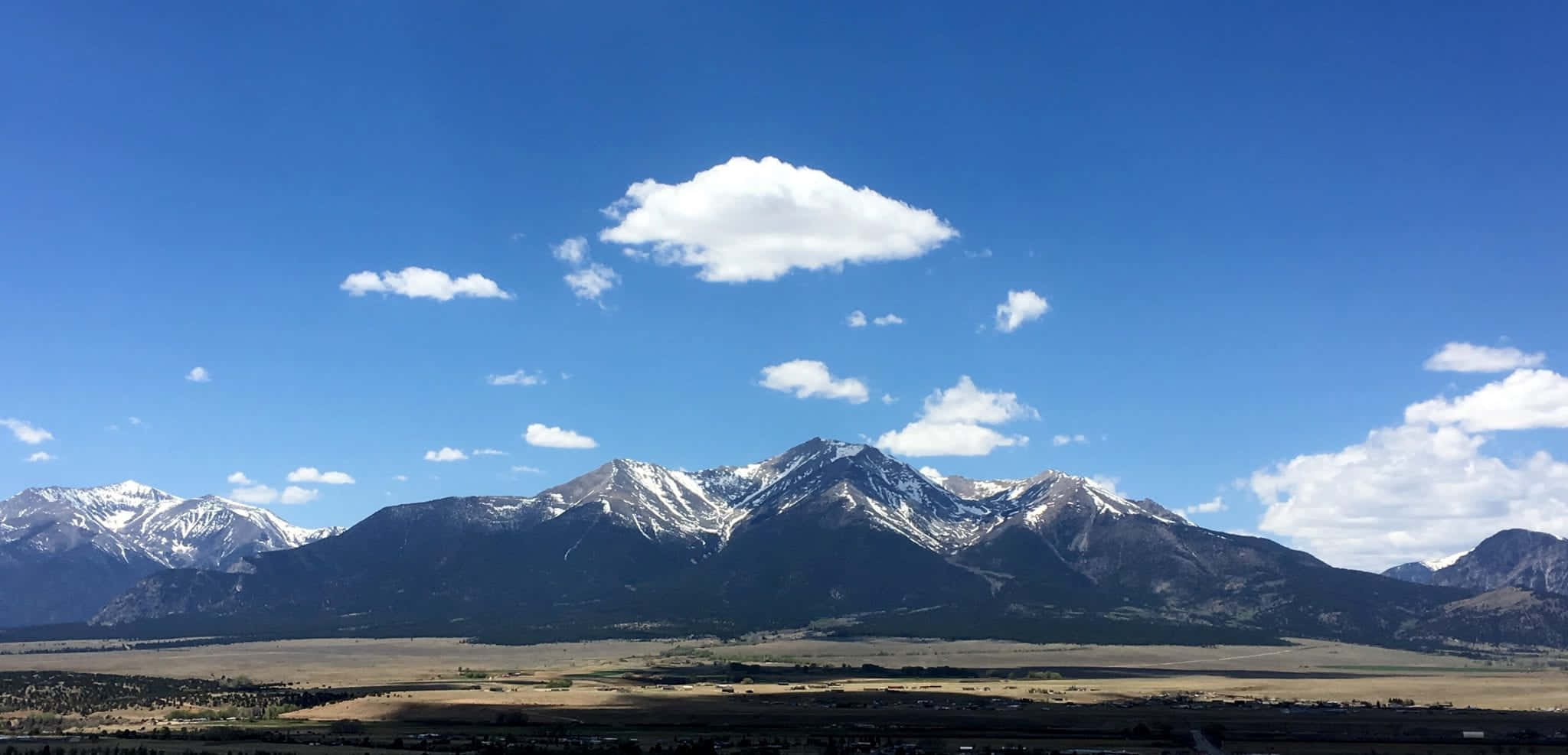 欣賞科羅拉多山脈的絕美風光 壁紙 — 白雪覆蓋的山峰在廣闊的藍天下雄偉地聳立，藍天上點綴著蓬鬆的白雲，下方是金色的山谷。