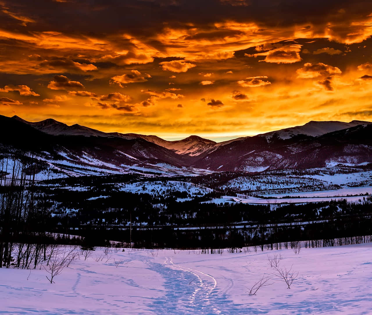 欣賞科羅拉多雄偉山脈的美景 壁紙 — 日落時分白雪覆蓋的山谷，暗黑山巒之上有火橙色雲彩，前景雪地上有蜿蜒的軌跡。