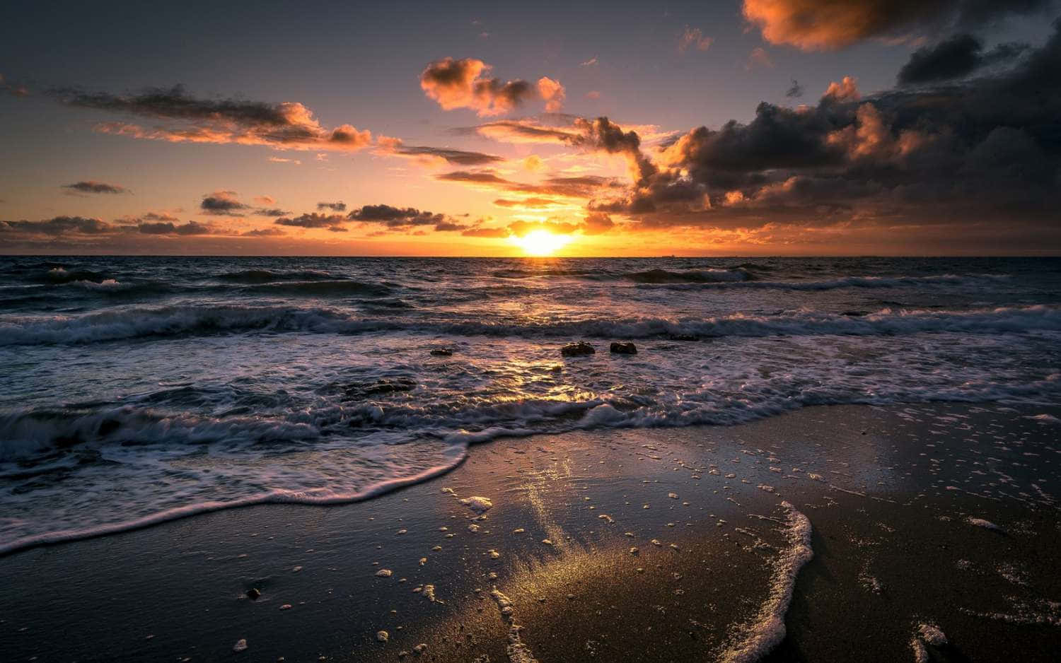 欣賞蔚藍大海與白沙海灘 壁紙 — 夕陽將天空染成火熱的橙色和紫色，海浪拍打著深色沙灘。