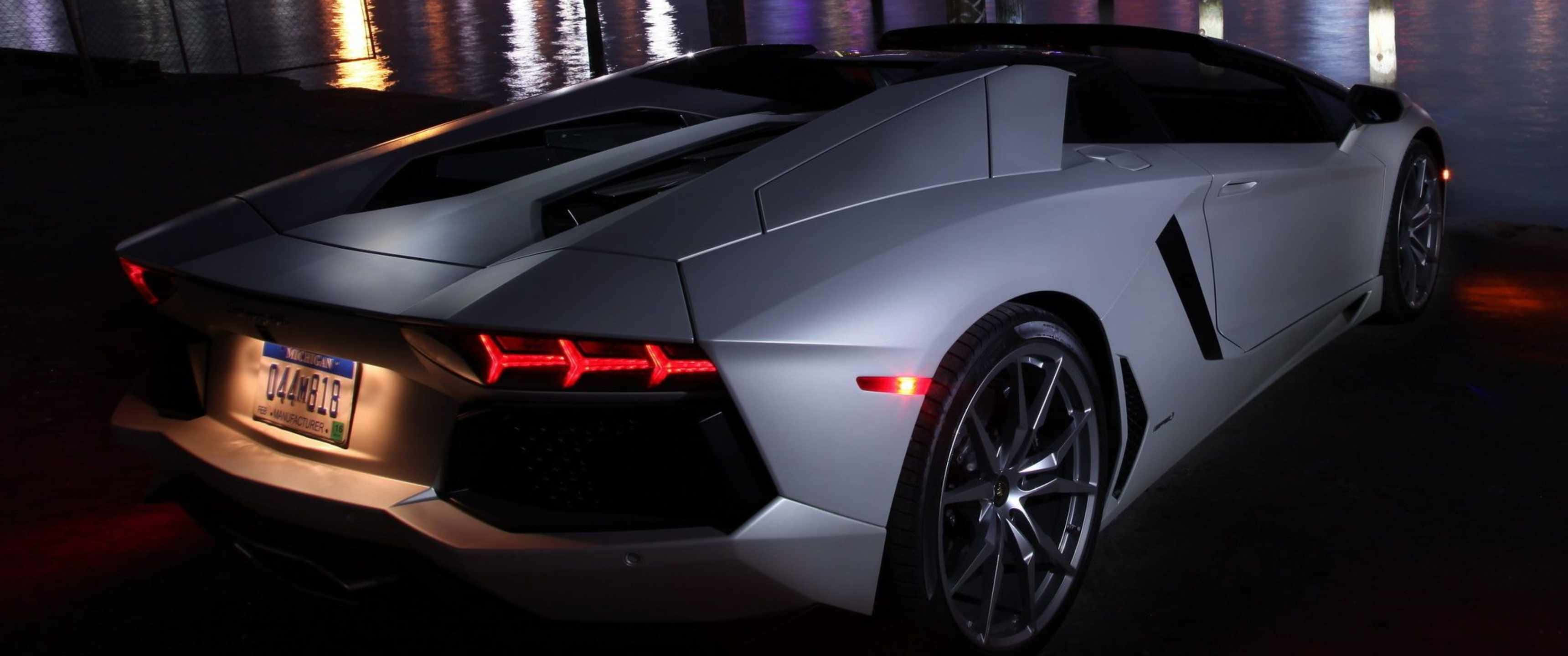 「欣賞這輛 3440x1440p 蘭博基尼！」 壁紙 — 一輛時尚的銀色林寶堅尼Aventador跑車在夜間霓虹燈下閃耀，在濕漉漉的路面上留下倒影。