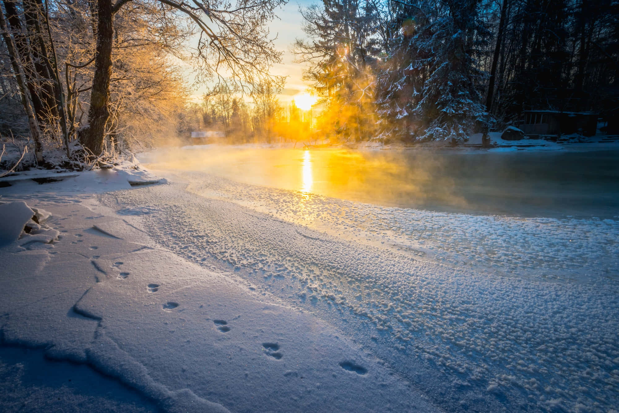 欣賞雪覆蓋的山巒之美 壁紙 — 陽光溫暖地照耀在一條被雪覆蓋的河流上，河流散發著蒸汽，腳印通向迷霧籠罩的地平線。